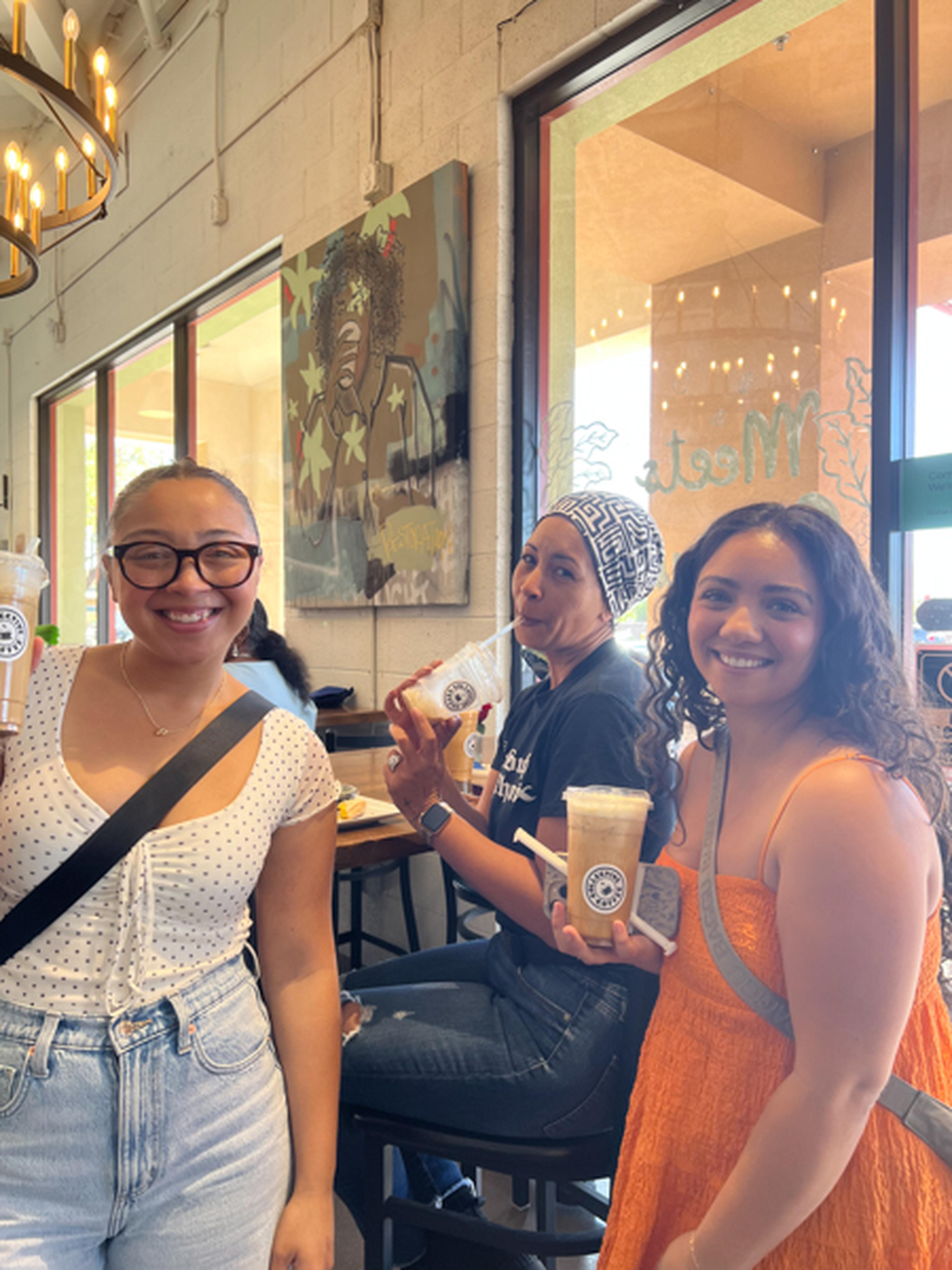 Three friends smile at Collective Coffee, two standing with drinks in hand while one sits at a table