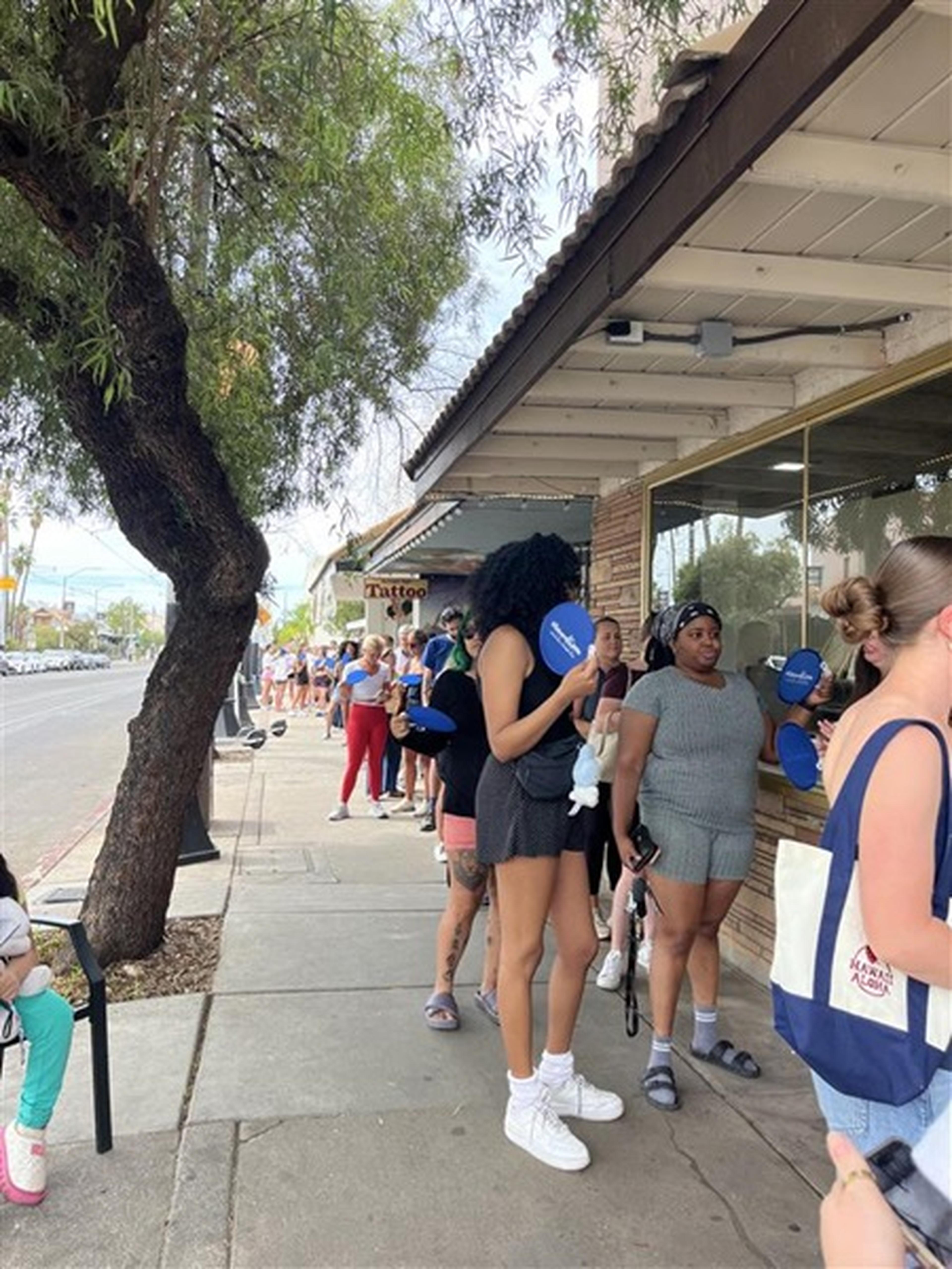 On a tree‑lined sidewalk, customers in casual summer attire wait outside the 1989 Bake House with fans in hand.