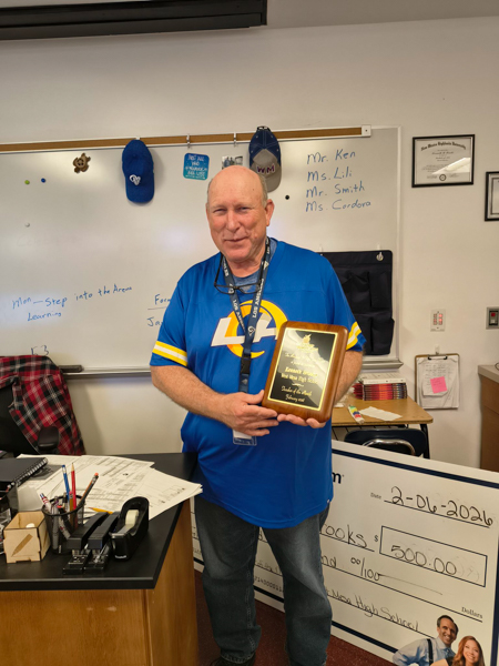 Mr. Brooks holds his teacher appreciation plaque in a classroom, standing near a desk with papers and a whiteboard in the background.