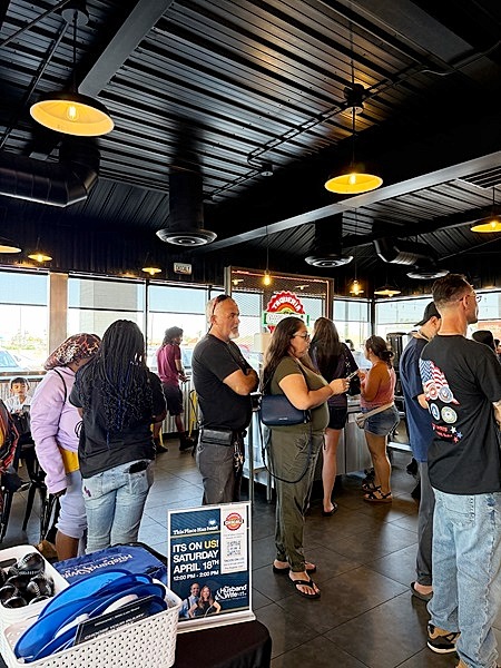People wait in line at Taqueria Chicali, surrounded by industrial lighting and large windows, while a table sign promotes an April 15th event.