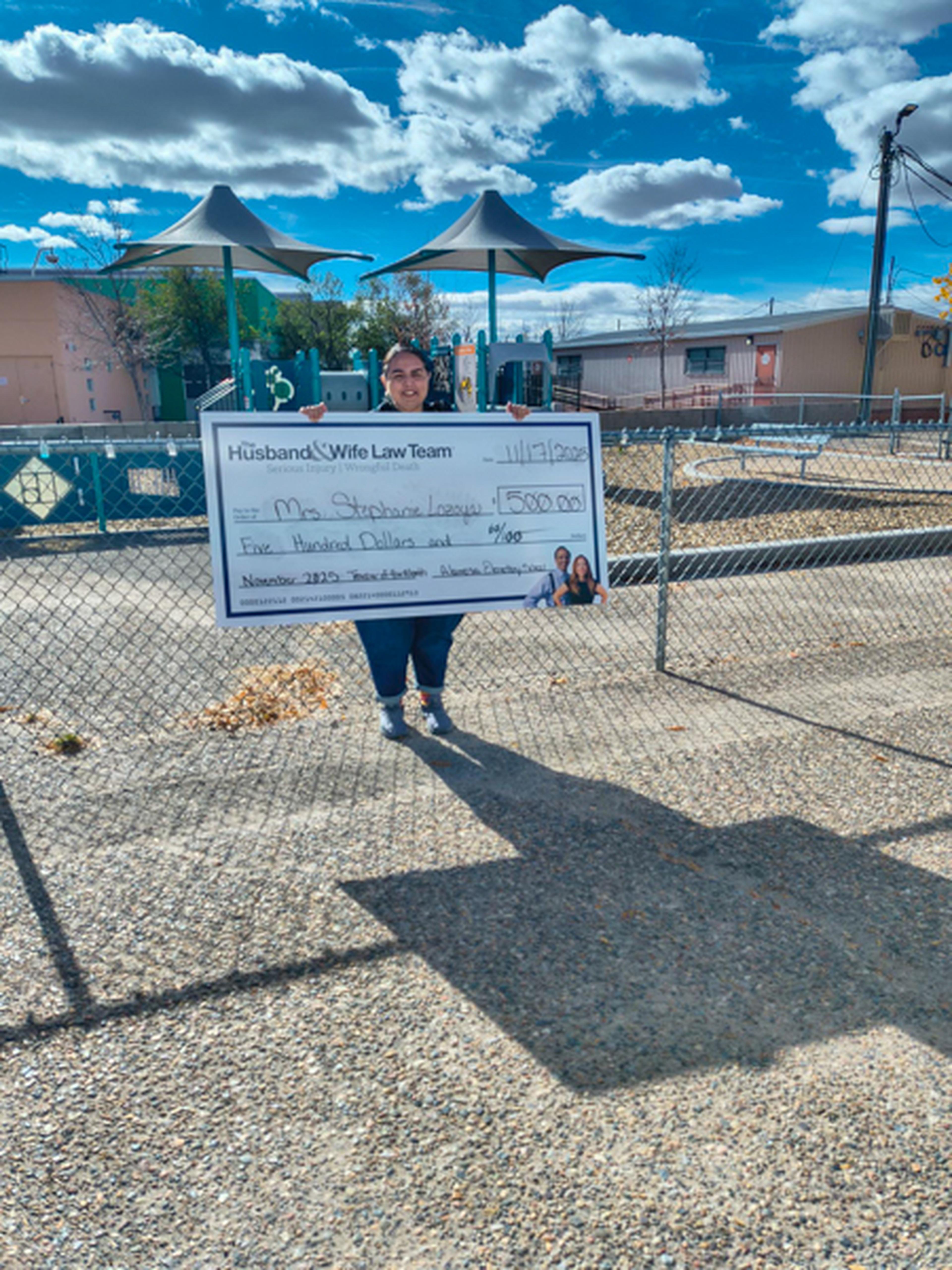 Teacher in front of school playground holding her oversized check for $500