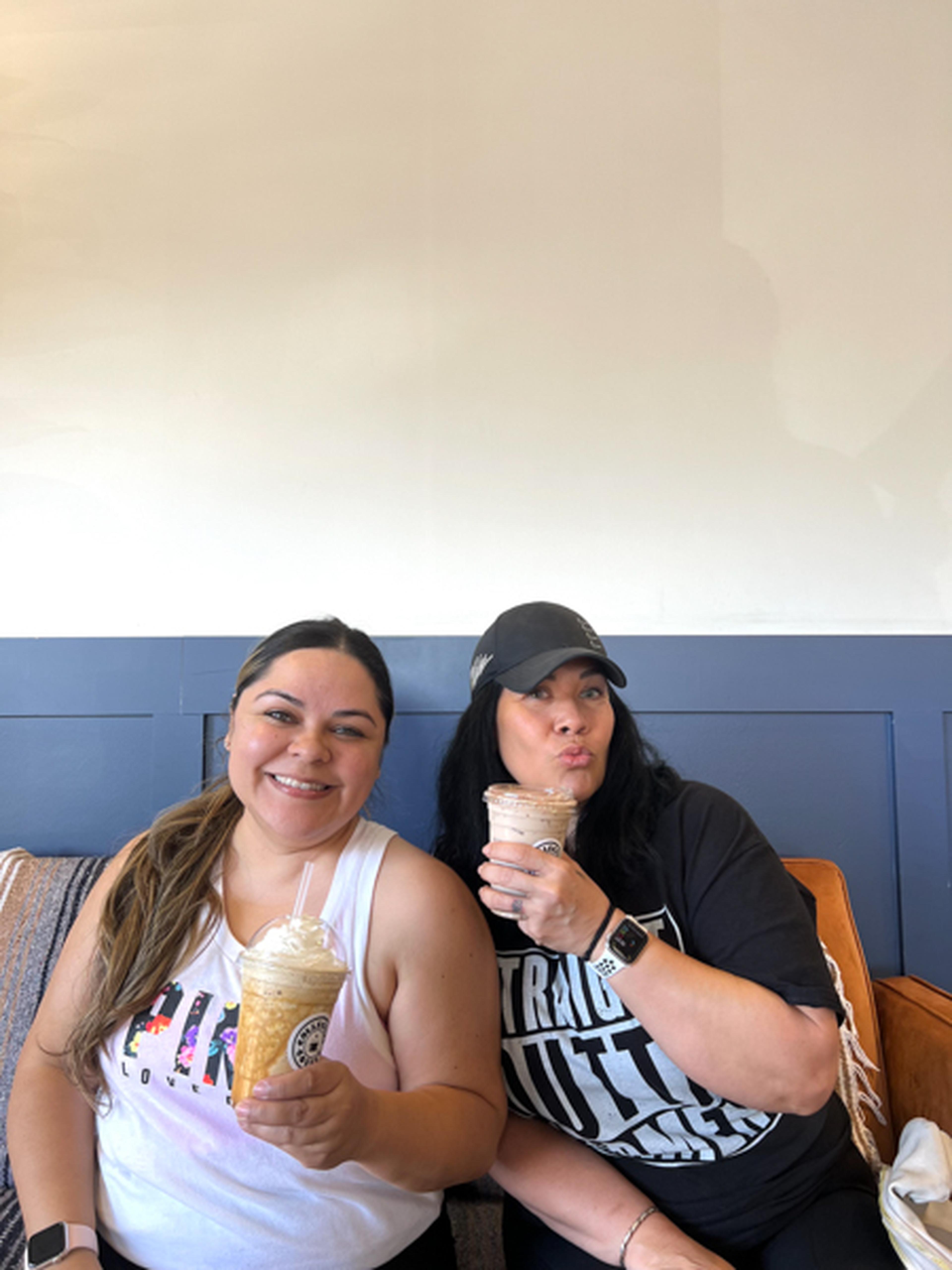 Two women relax on a couch with iced coffees, smiling warmly.