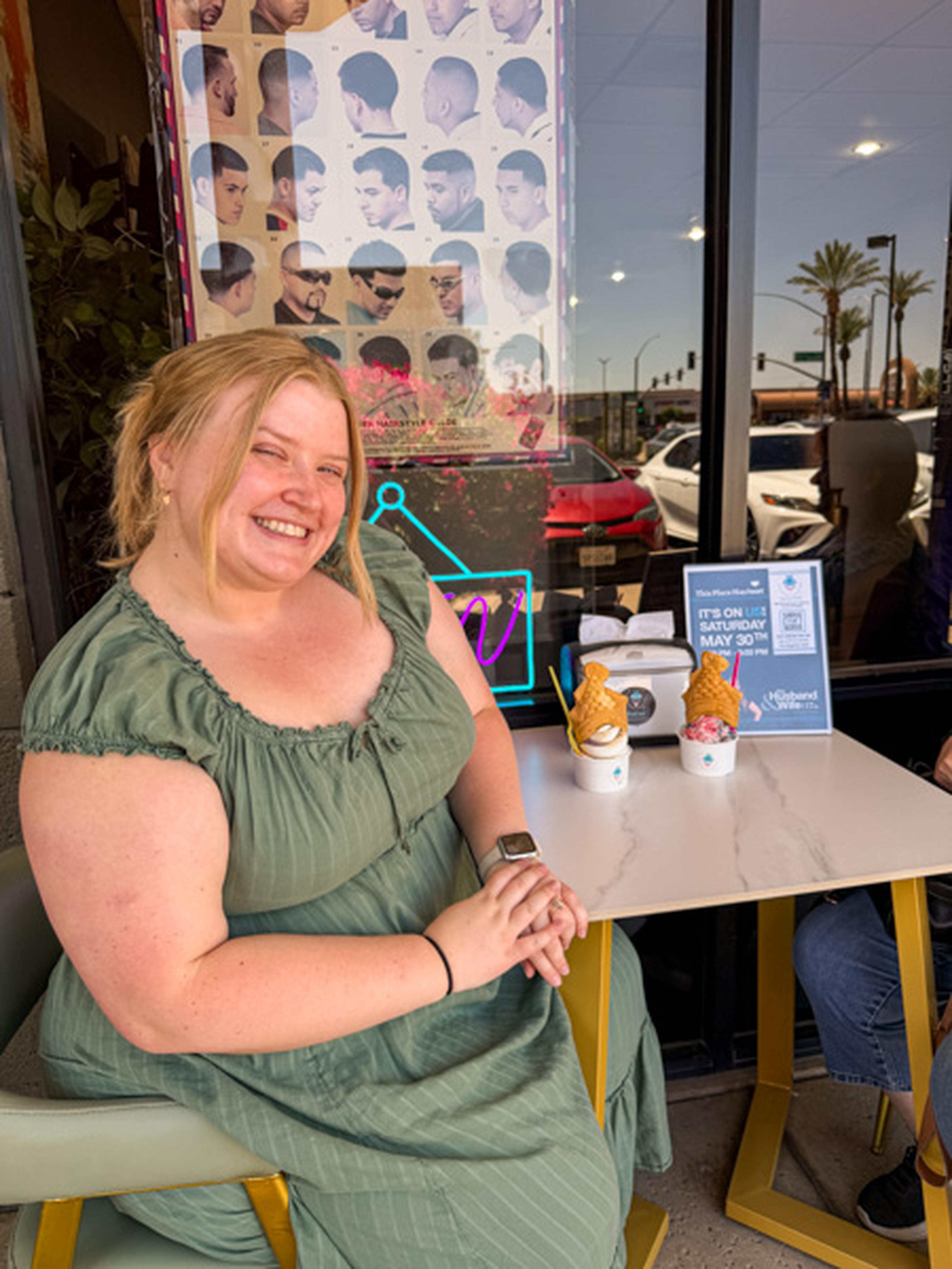 A woman in a green dress smiles at an outdoor table topped with ice cream cones, with a neon sign and hairstyle poster glowing in the background.