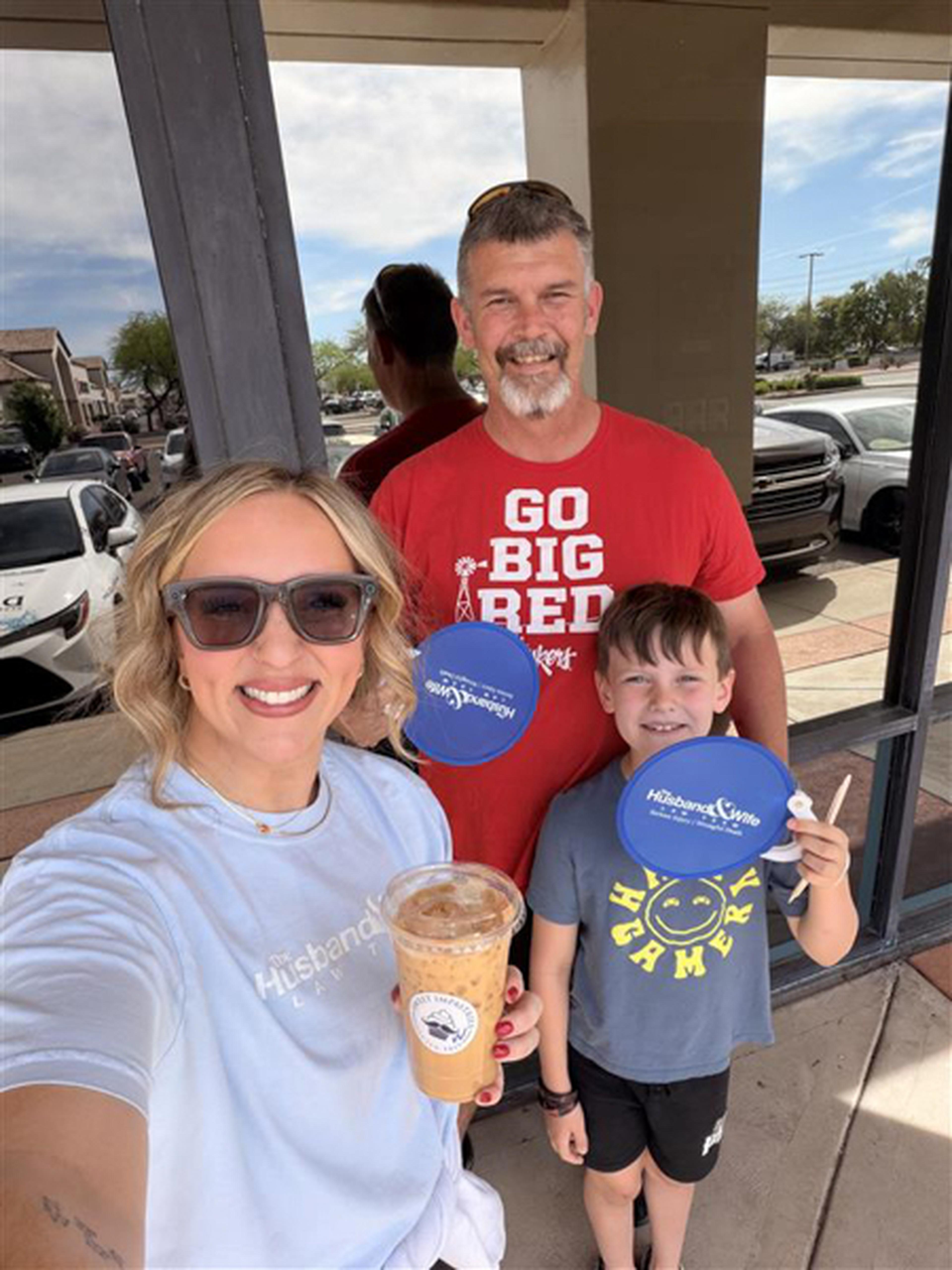 Jen holds a drink while posing outside Sweet Impastries in Gilbert. Background includes a man in a red shirt and a boy holding a blue fan disk.