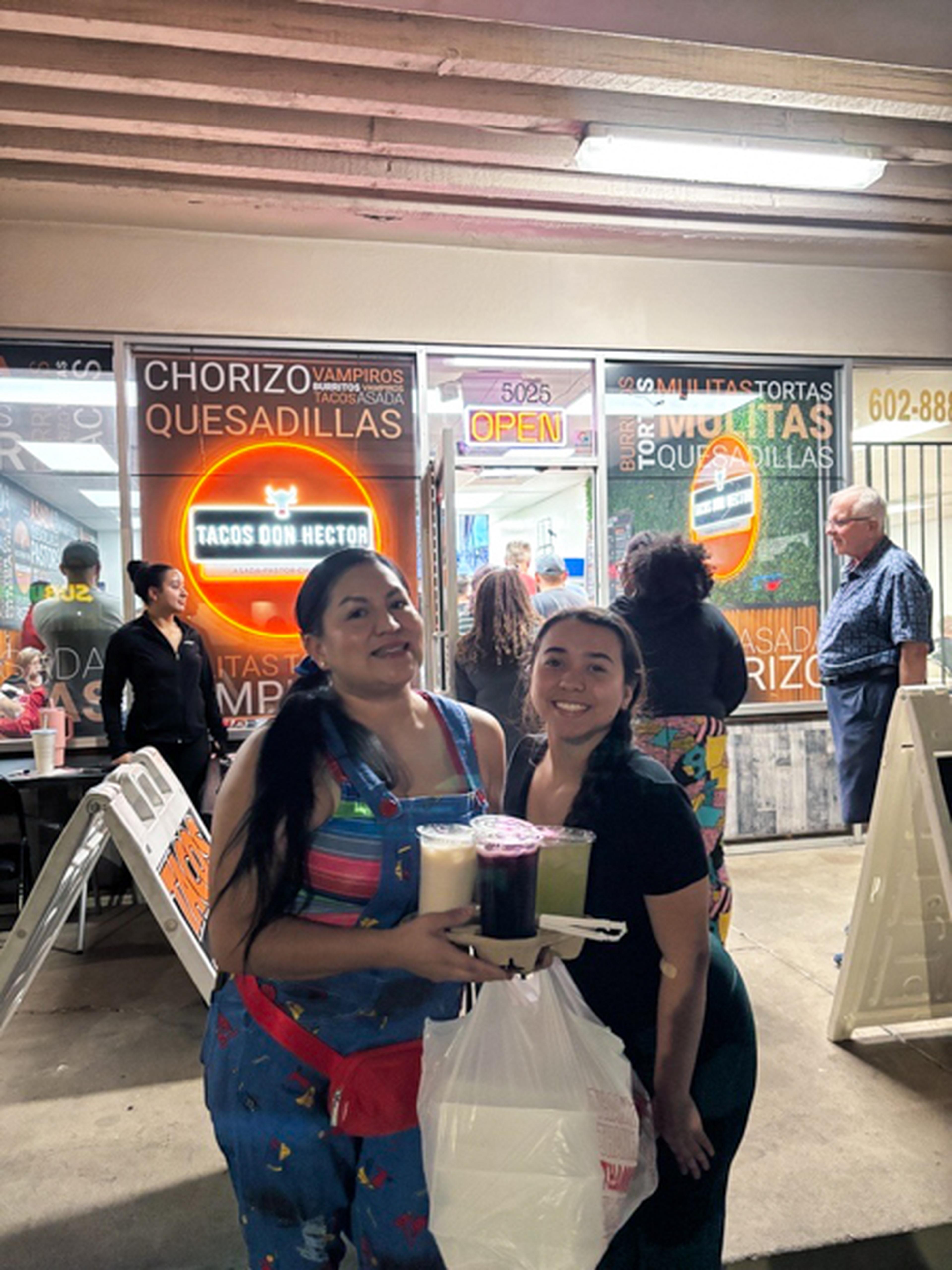 Two women smiling and holding drinks outside a taco shop at night.