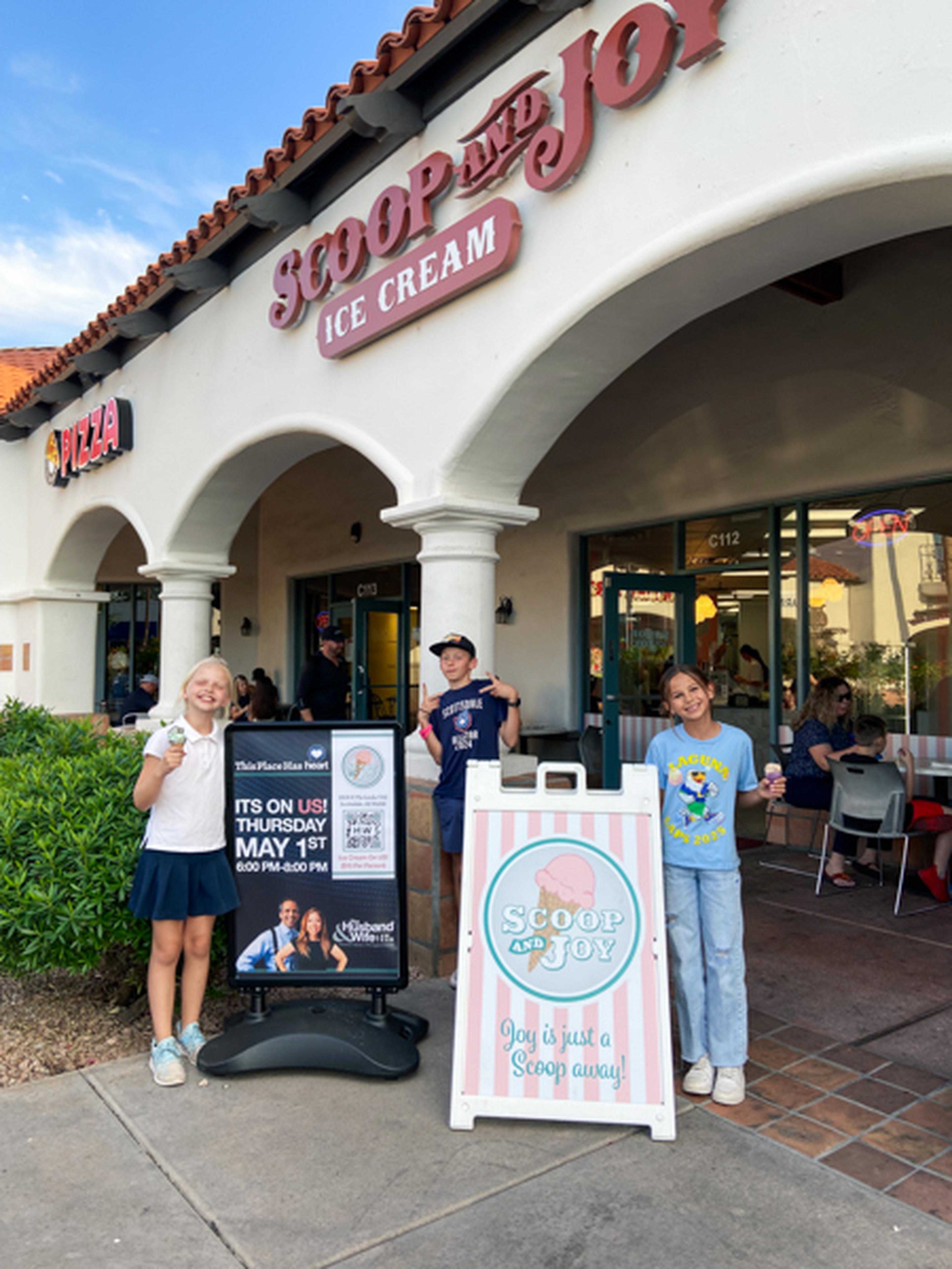 Three people stand outside "Scoop and Joy" ice cream shop, smiling near signs.