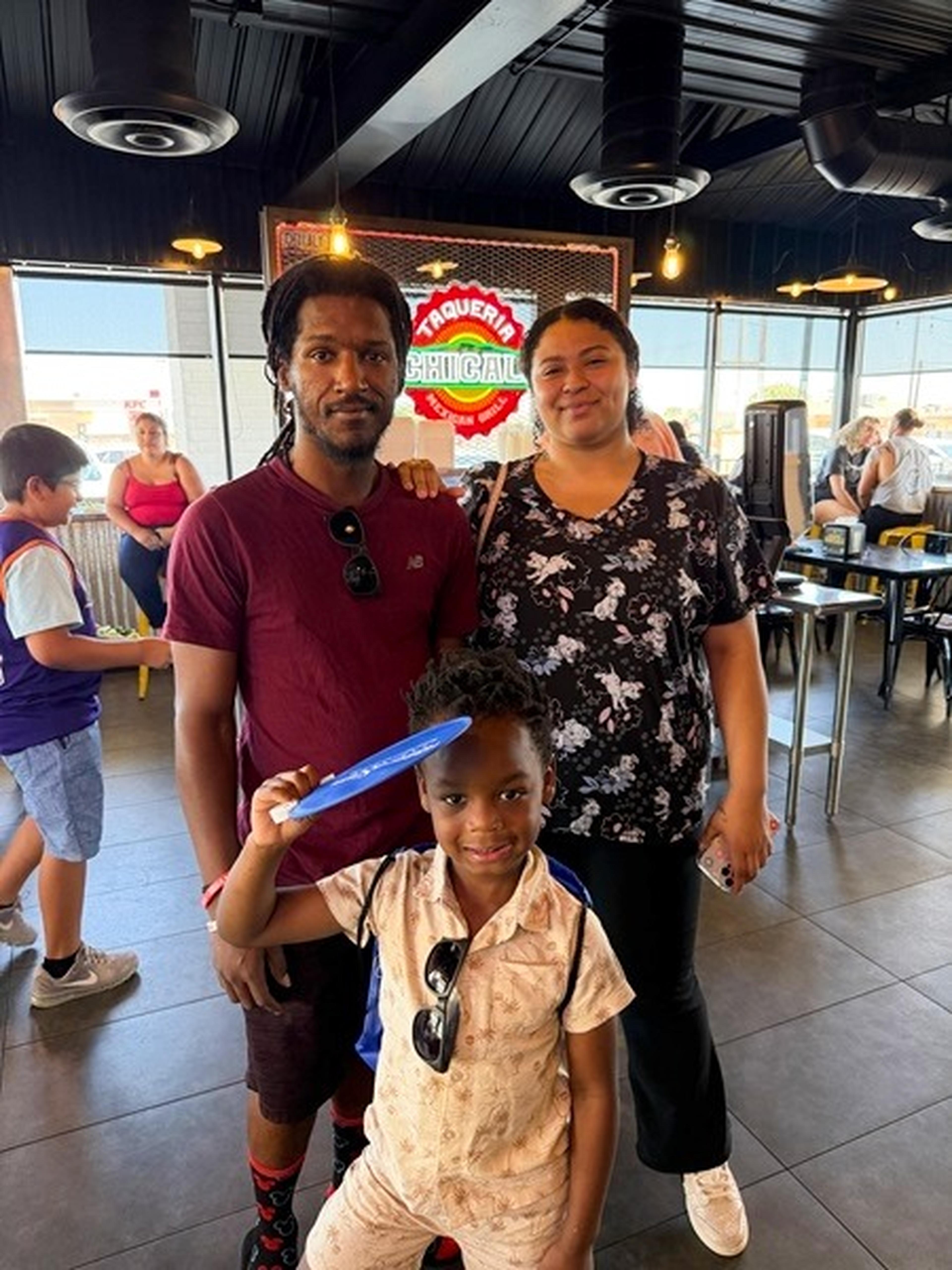 A family of three poses inside the restaurant, with a colorful sign behind them and the child holding a blue fan