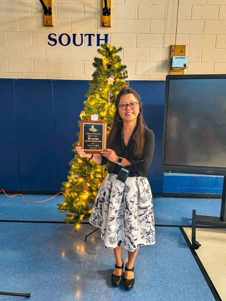 A woman in a black top and patterned skirt holds a plaque, standing beside a decorated Christmas tree.