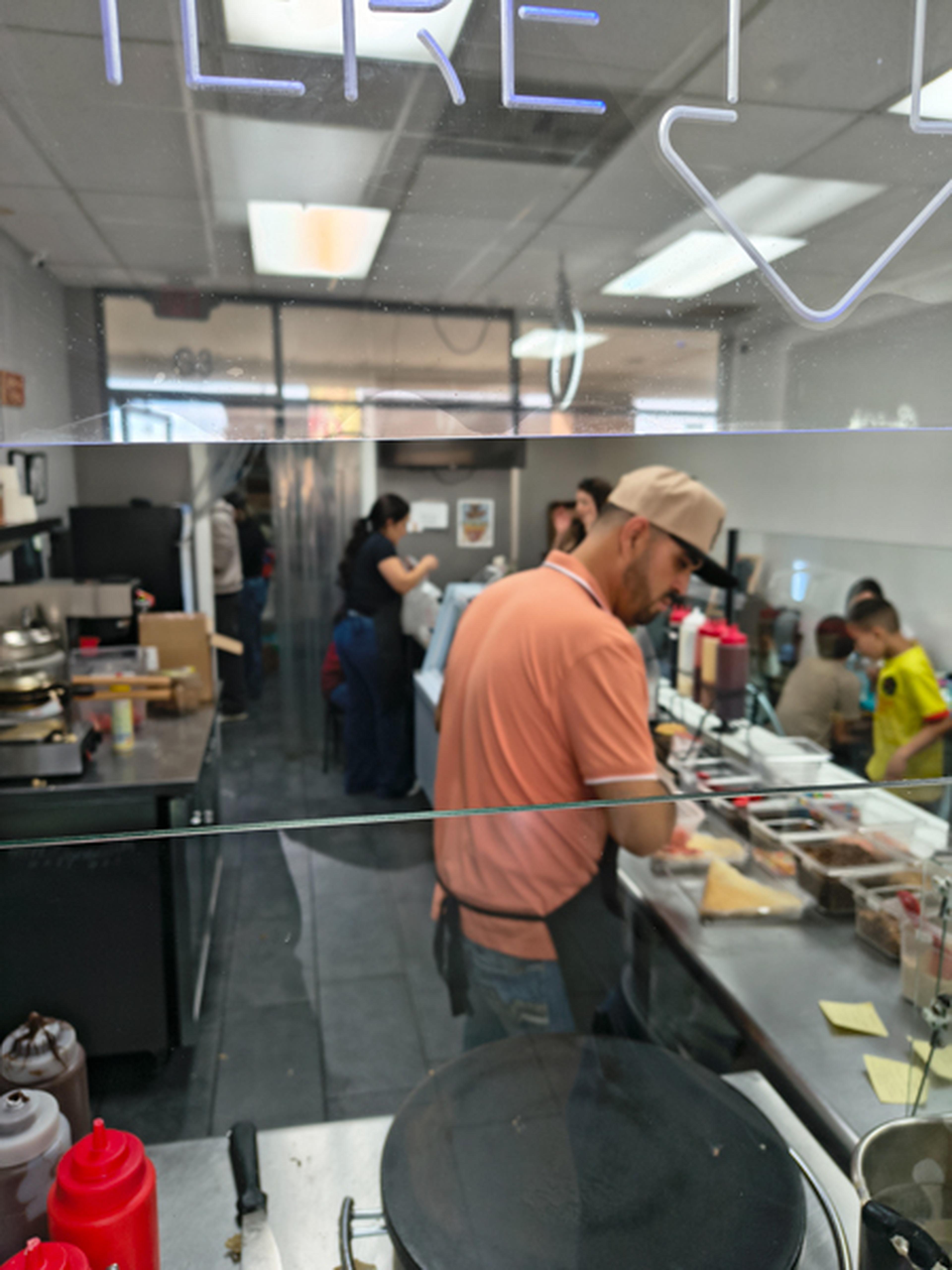 A man in an orange shirt and hat works Crepes and Waffles preparing food alongside several other cooks.