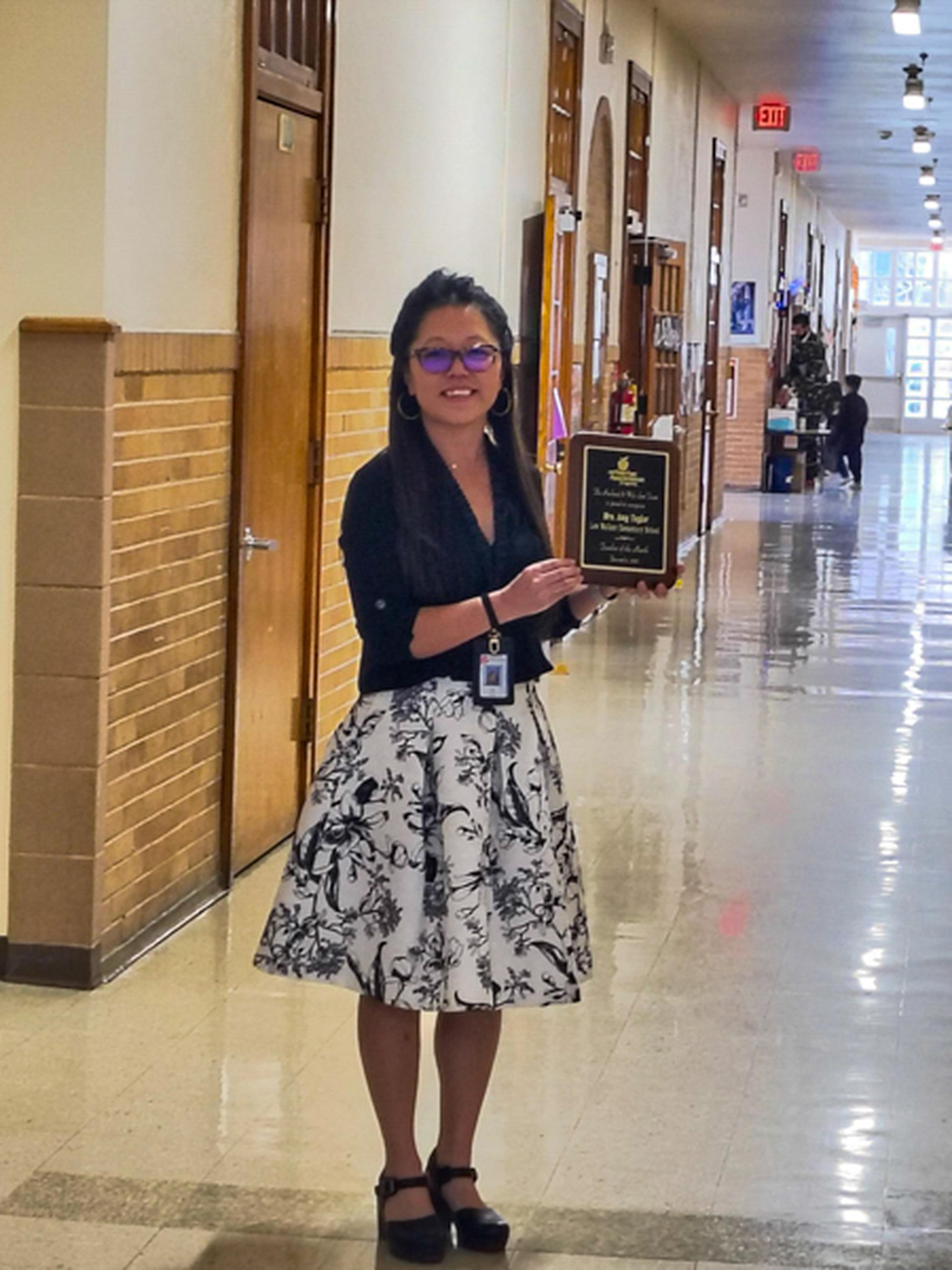 Woman in a blouse and floral skirt holds an award plaque, standing in a school hallway with a polished floor and open doors.