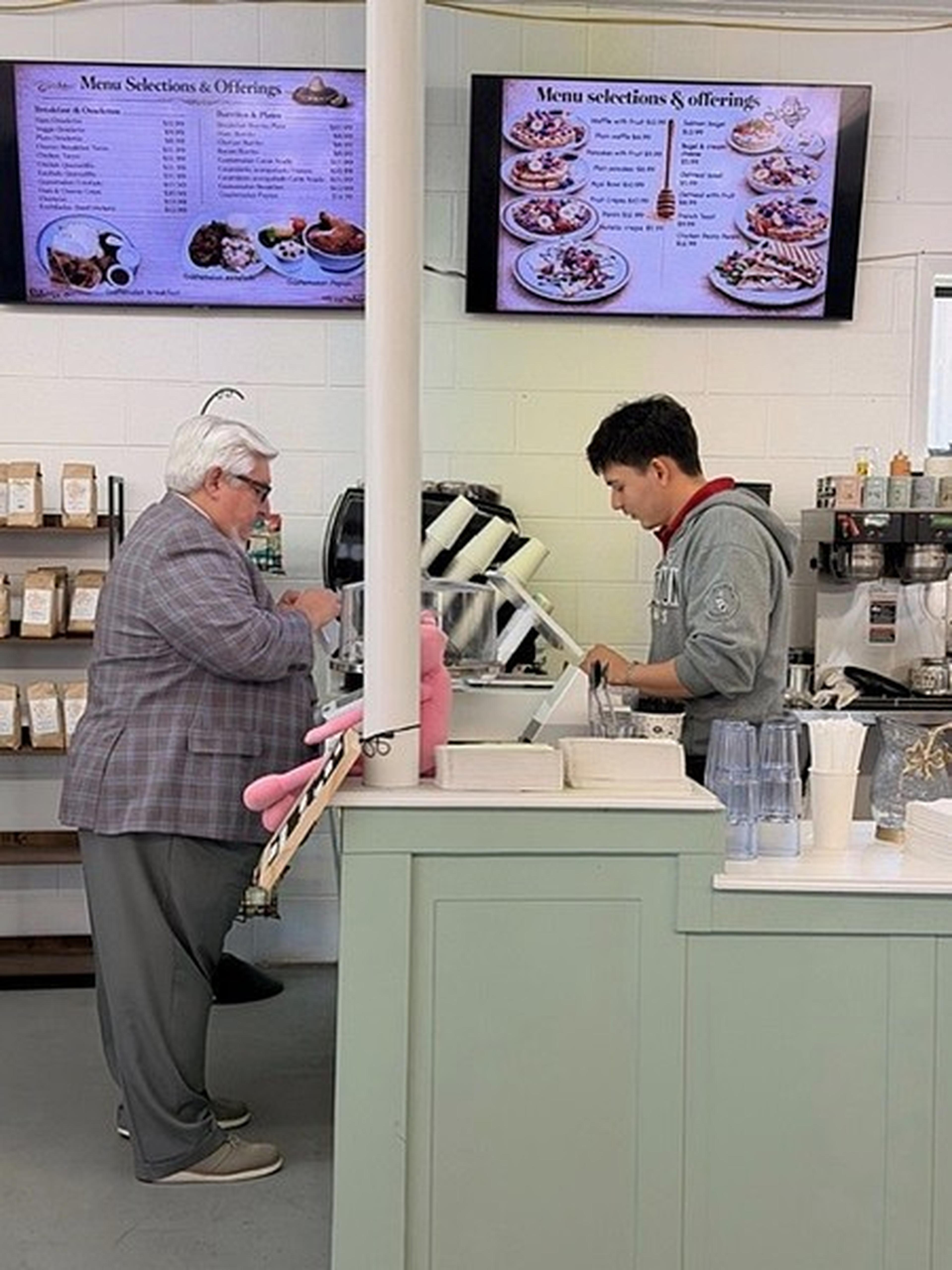 A Milk and Honey Caffe customer chats with the barista at the counter, framed by glowing menu screens and coffee