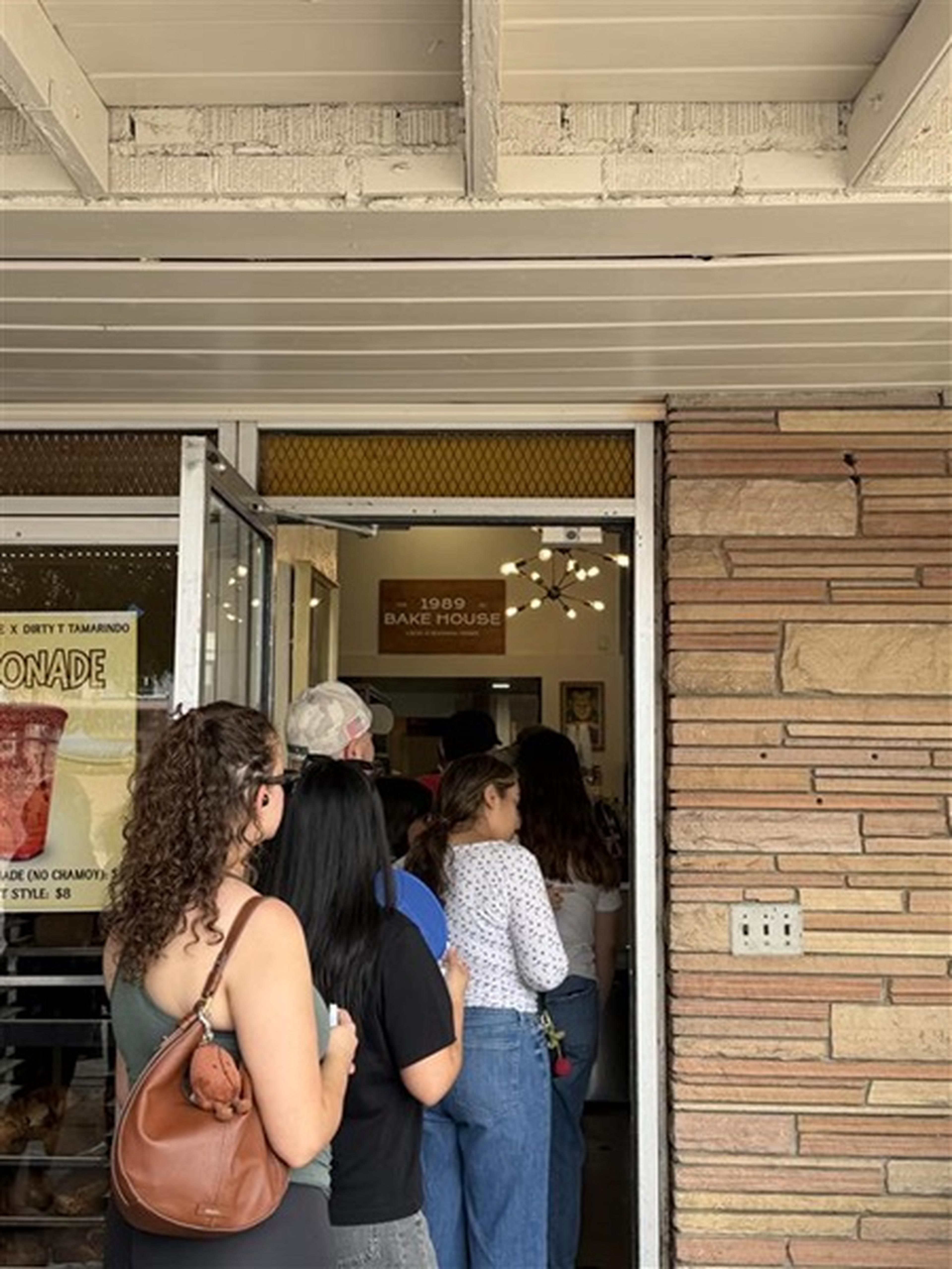 People wait in line outside the 1989 Bake House, with a lemonade poster visible on the glass door.