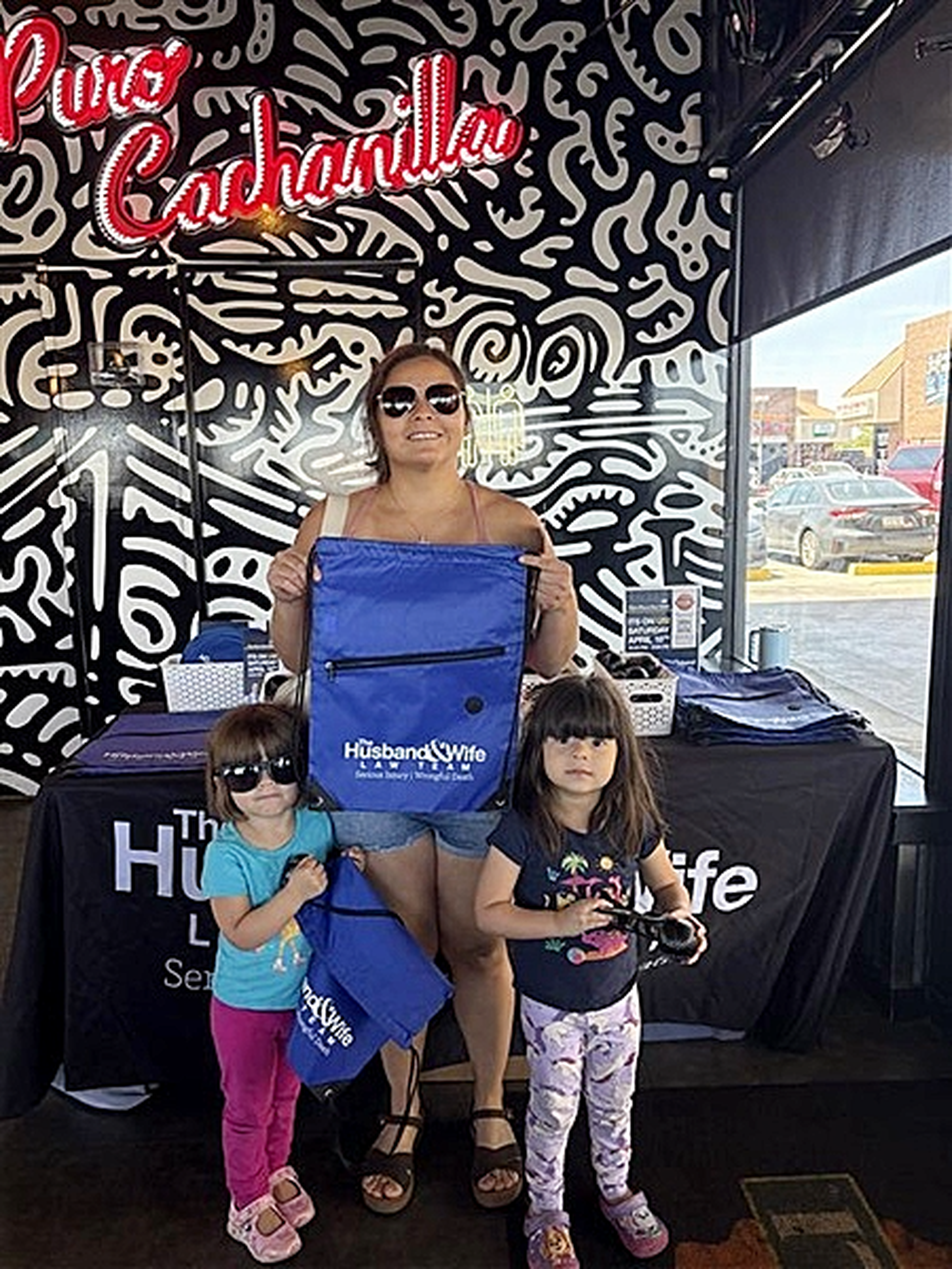 Inside Taqueria Chicali, a woman and her two children pose with blue backpacks beside a display table and bold black‑and‑white wall art.