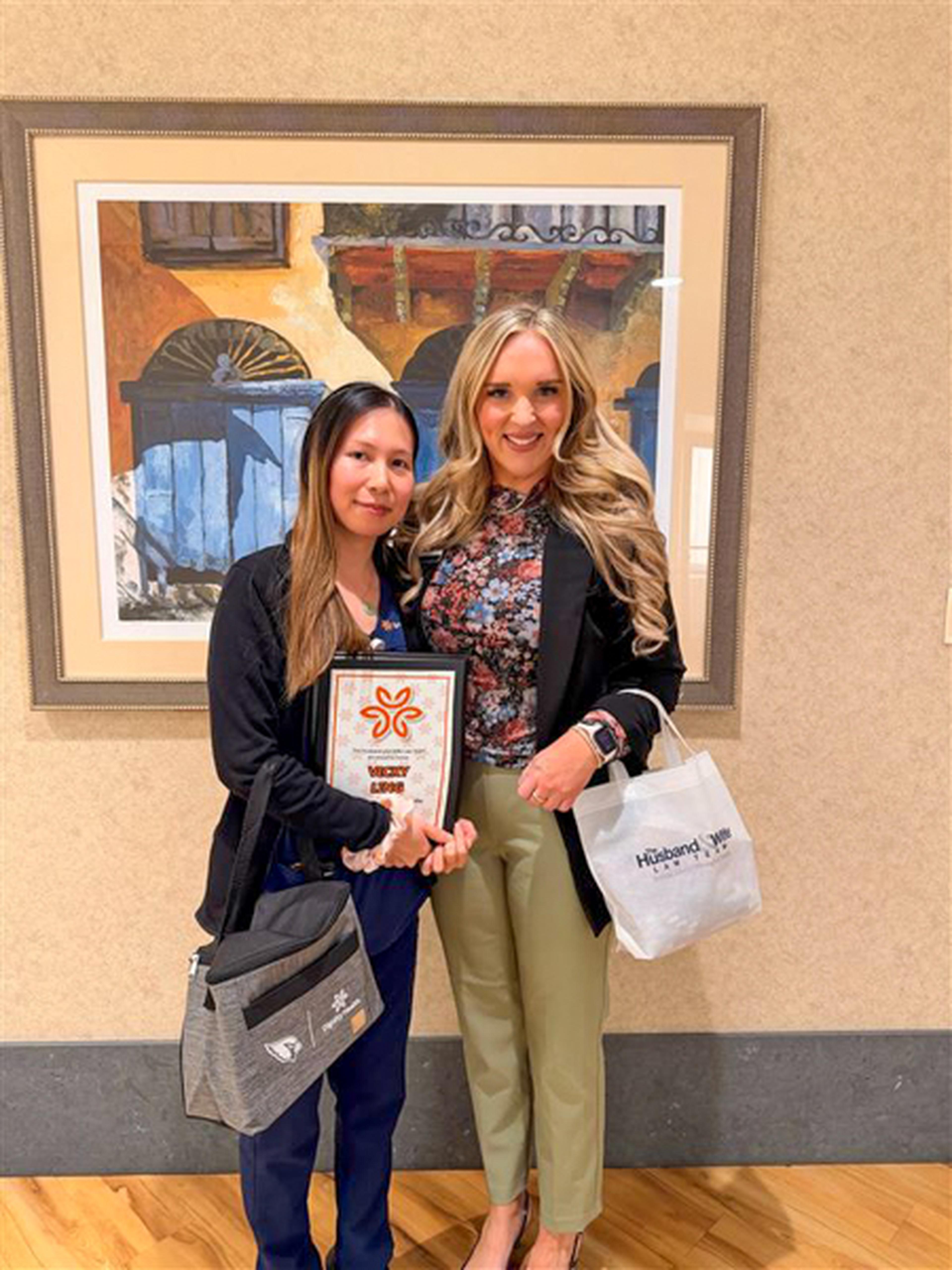 Jen form the Husband & Wife Law Team  and Vicky with a plaque and gift bags, smiling in front of framed artwork.