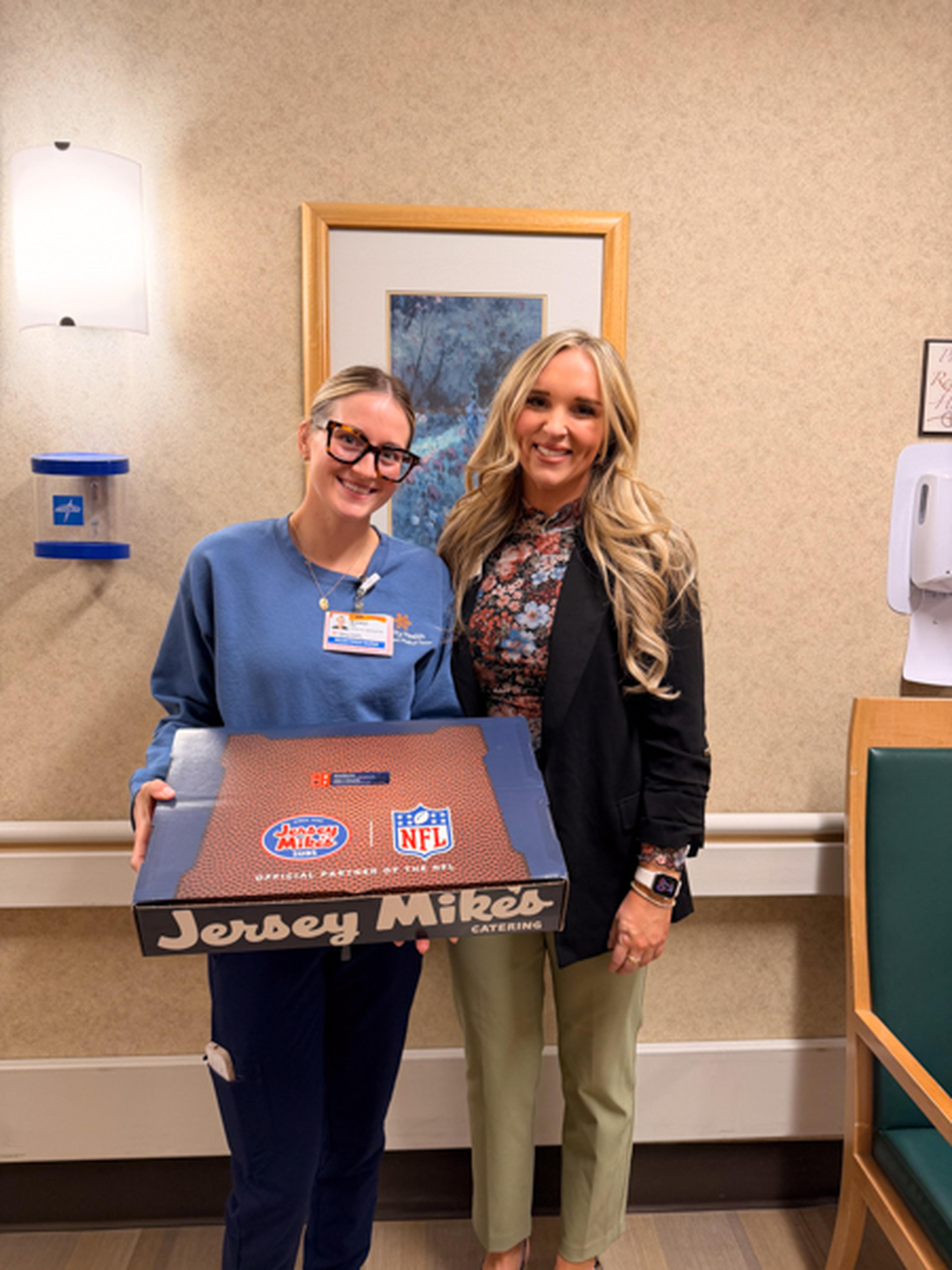 Jenn smiling indoors; one in scrubs holds a Jersey Mike's catering box. Framed art and medical equipment visible in the background.
