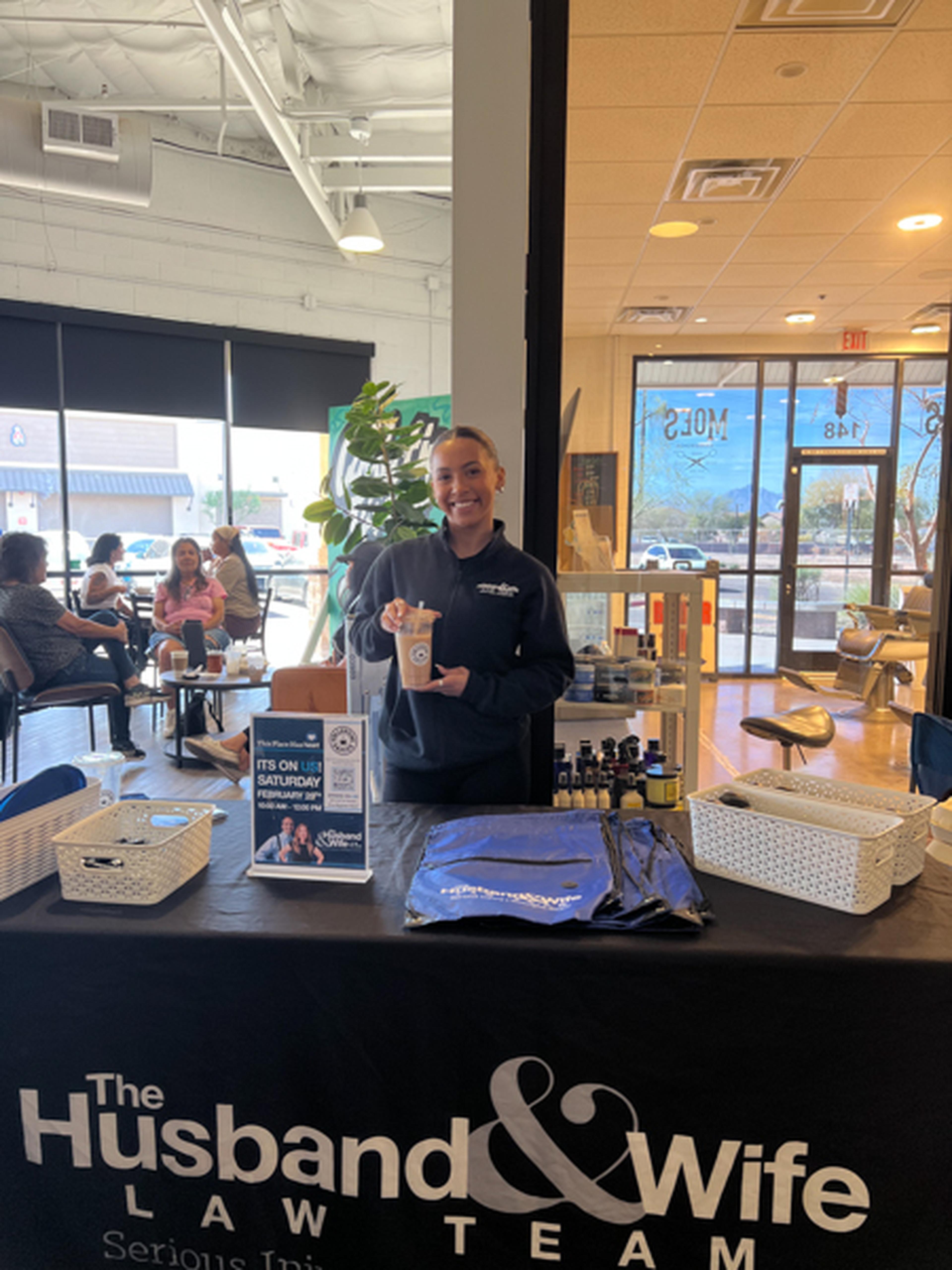 Hannah stands smiling behind a promotional table for "The Husband & Wife Law Team" inside a cafe, with brochures and branded items displayed.