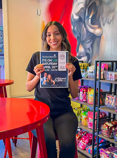 Smiling woman holding an event poster inside Pop-Pops, a vibrant cafe with colorful wall art and shelves of snacks in the background.