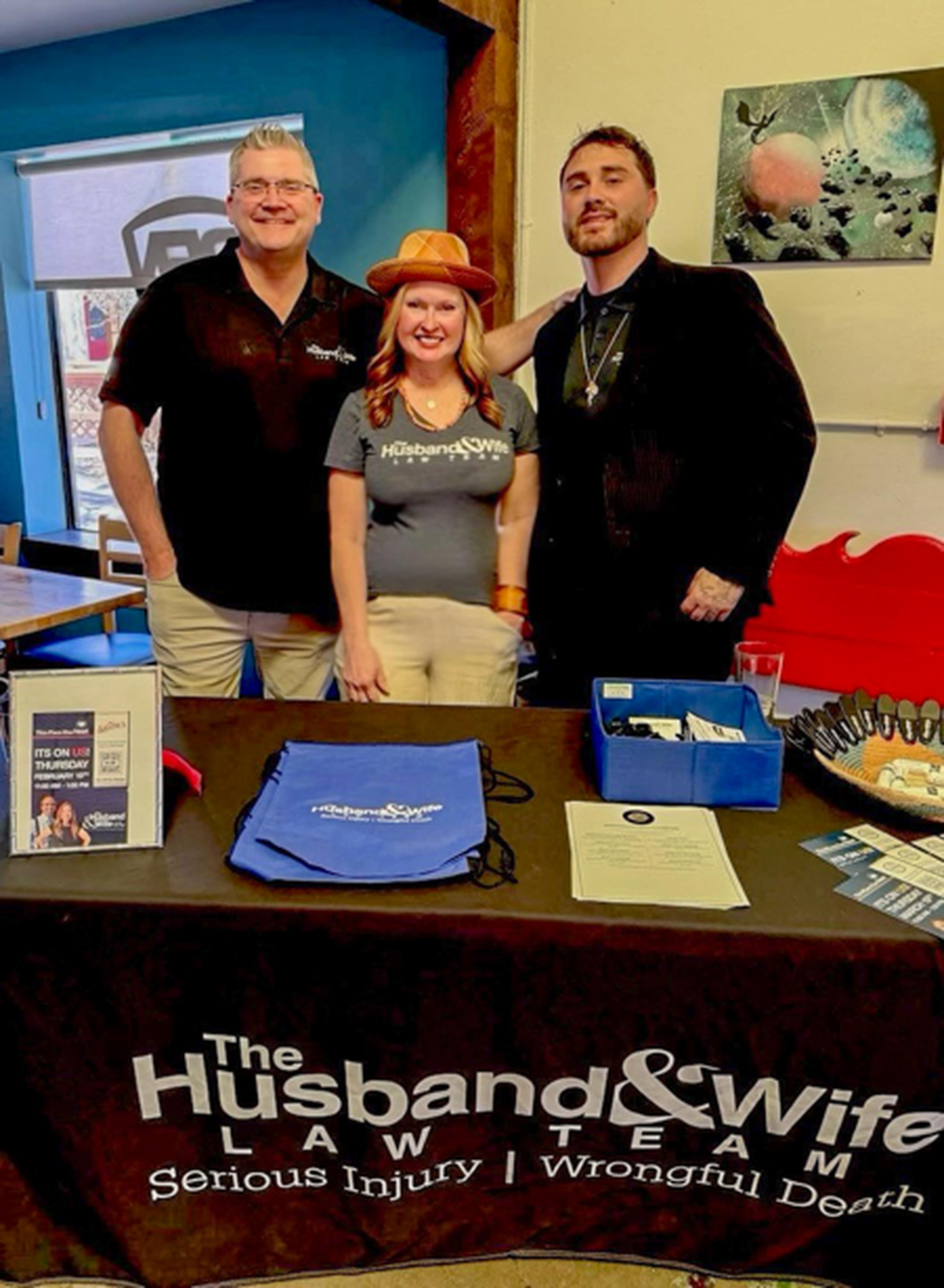 Brian, Lisa and Josh all stand behind a table with "The Husband & Wife Law Team" banner, brochures, and merchandise.