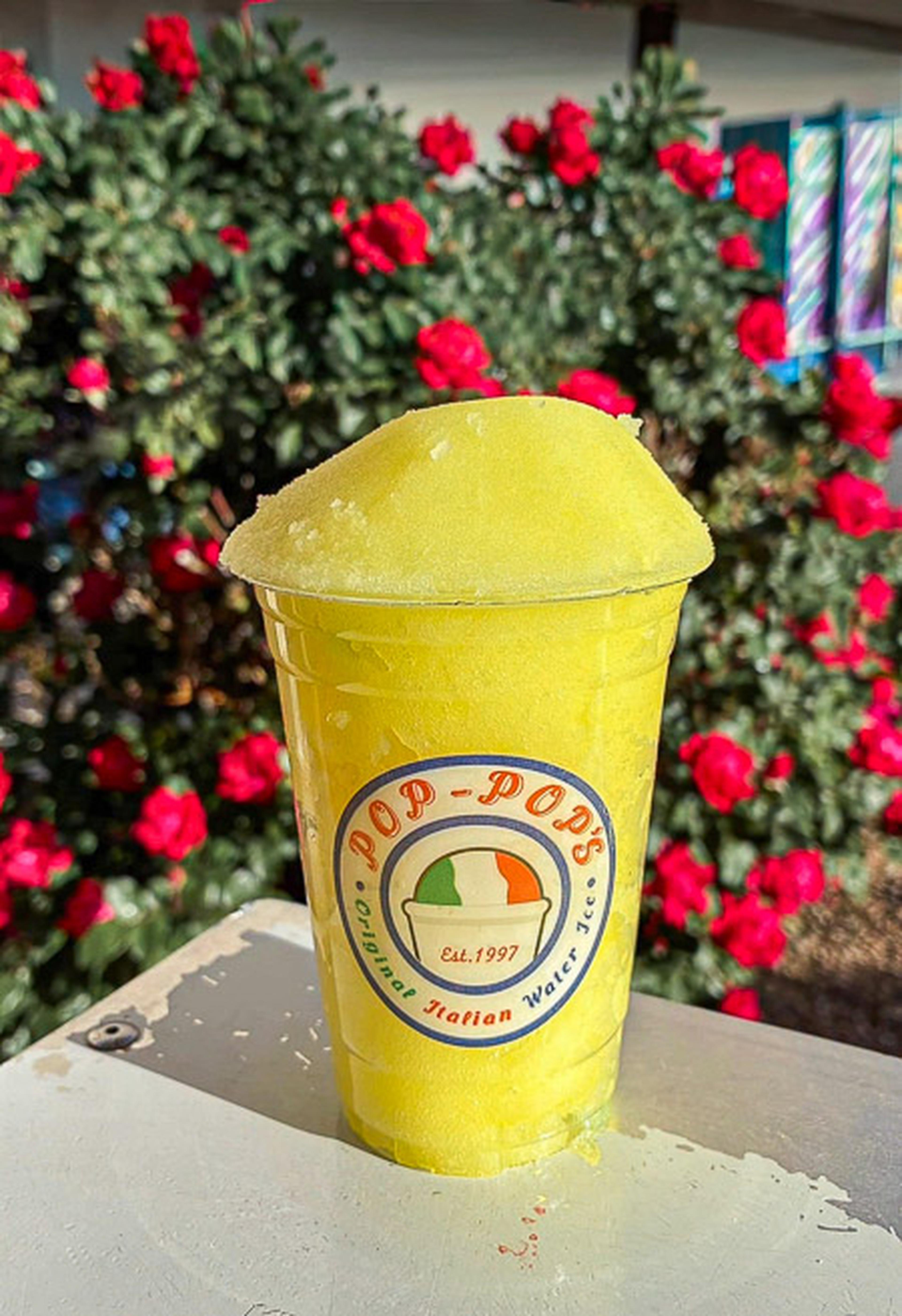 A bright yellow Italian ice in a Pop-Pop's branded cup on a table, with red flowers and greenery in the background.