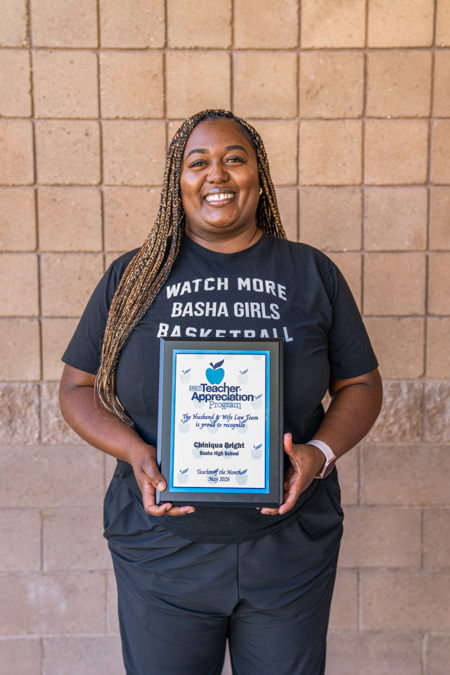 Teacher Appreciation spotlight as Coach Bright poses with her plaque, his “Watch More Basha Girls Basketball” shirt popping against the brick wall behind him. Coach Bright stands proud holding her Teacher Appreciation plaque while sporting a “Watch More Basha Girls Basketball” shirt against a bold brick‑wall backdrop.
