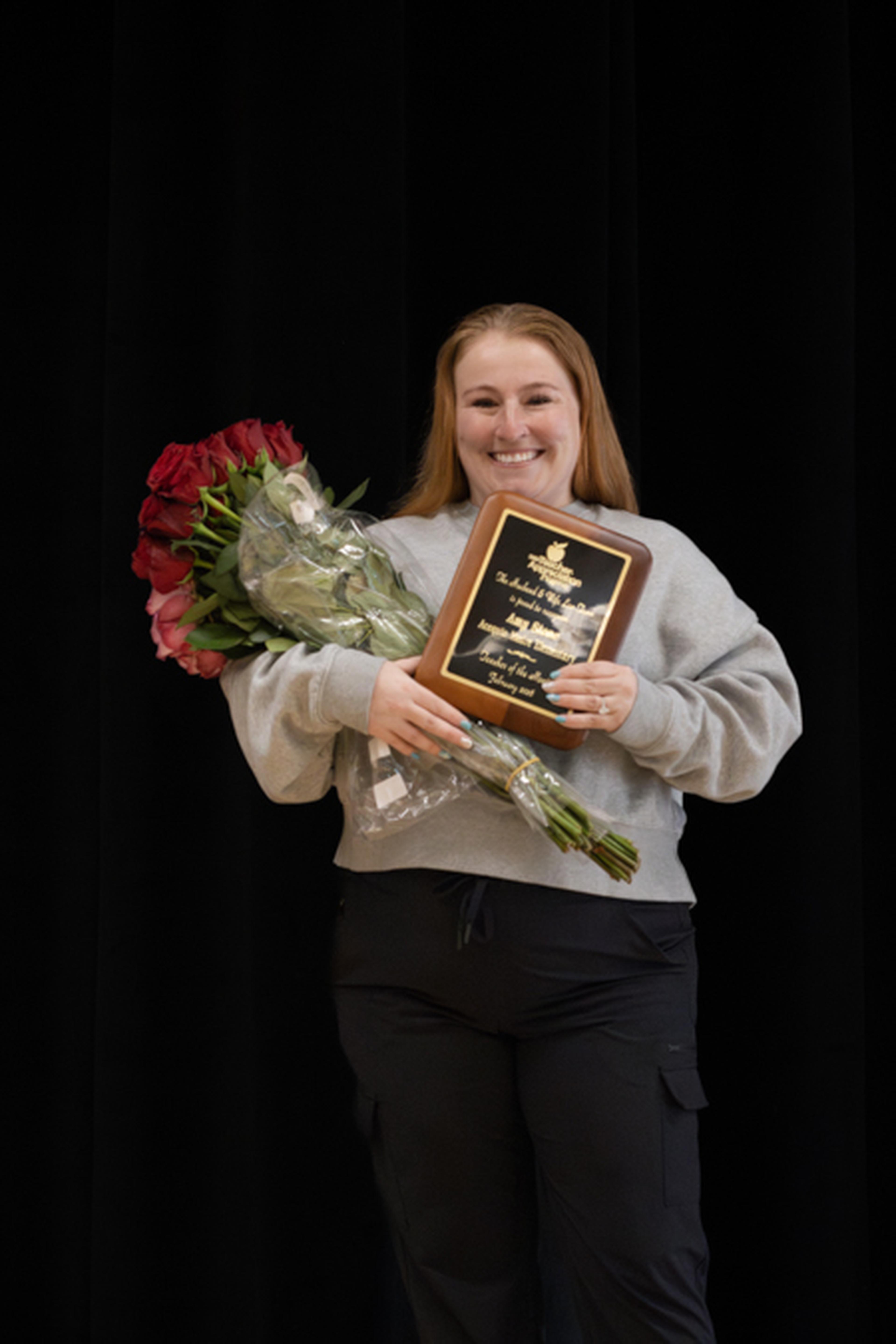 Ms. Amy smiles against a black backdrop, holding a bouquet of red roses and a plaque recognizing her teaching achievements.