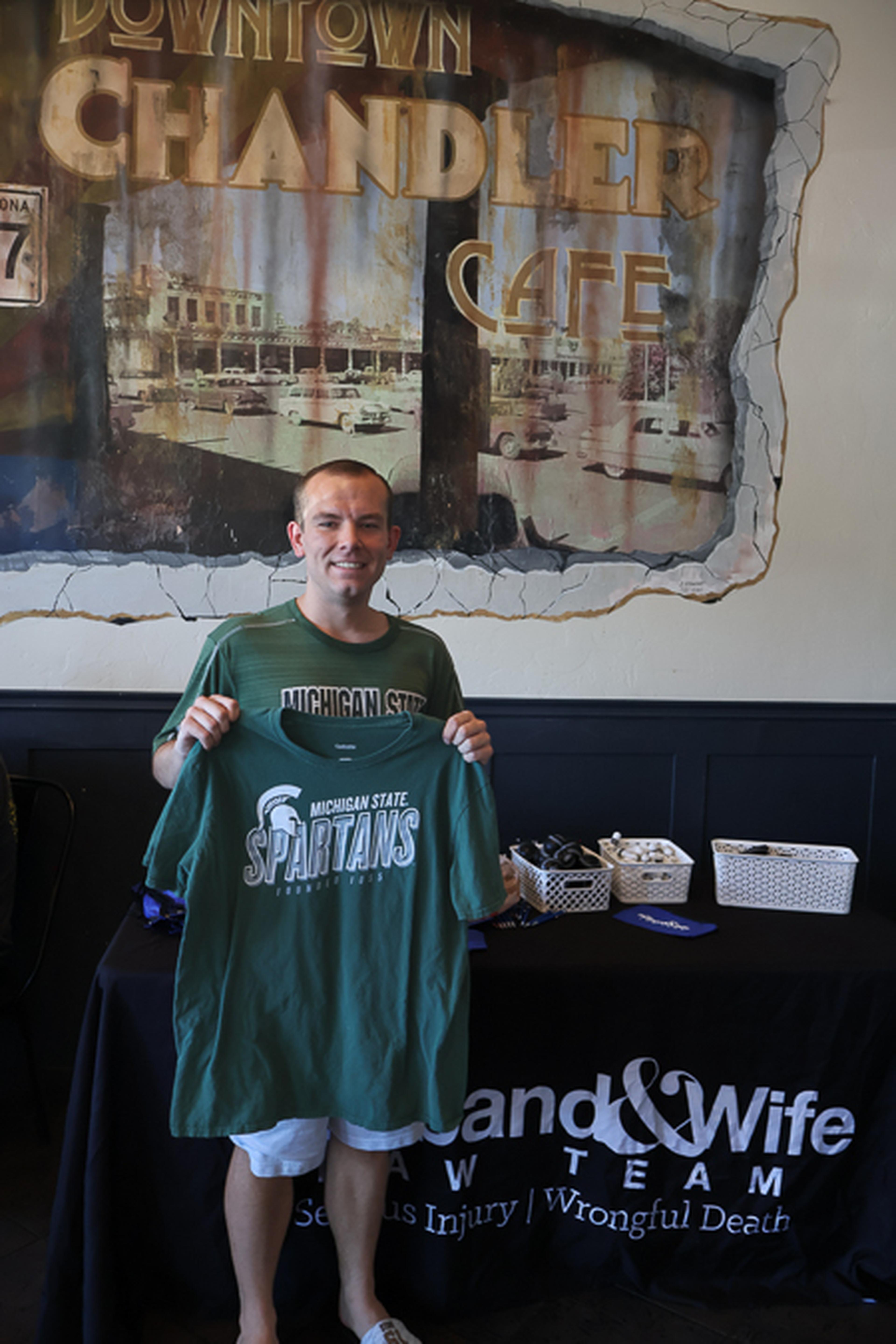 A community member shows off a Michigan State Spartans T-shirt at a Husband & Wife Law Team event, highlighting a shared connection to Mark Breyer's alma mater.