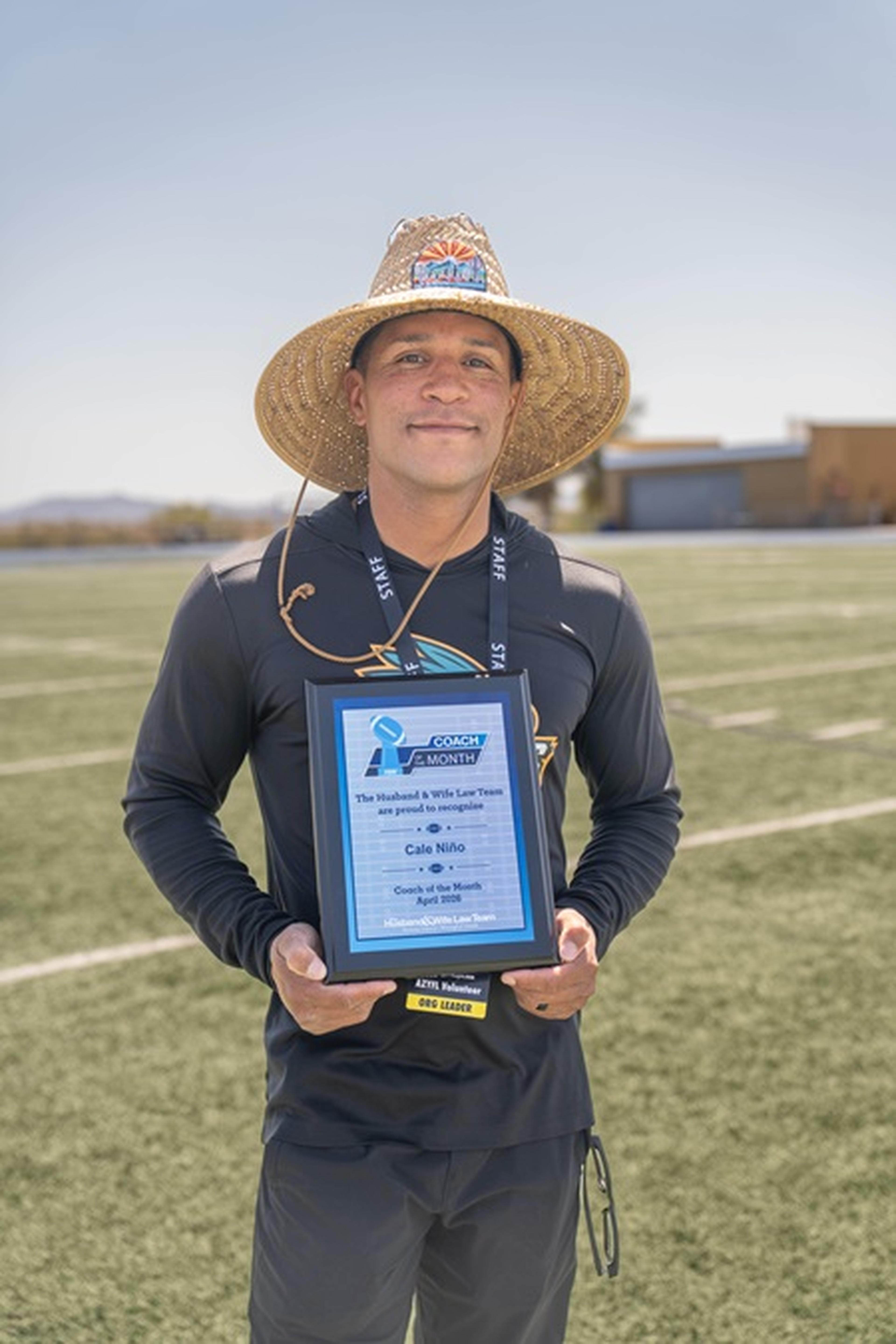 Coach Nino stands on the field holding a framed AYFL Coach of the Month award, dressed in a dark long‑sleeve shirt.