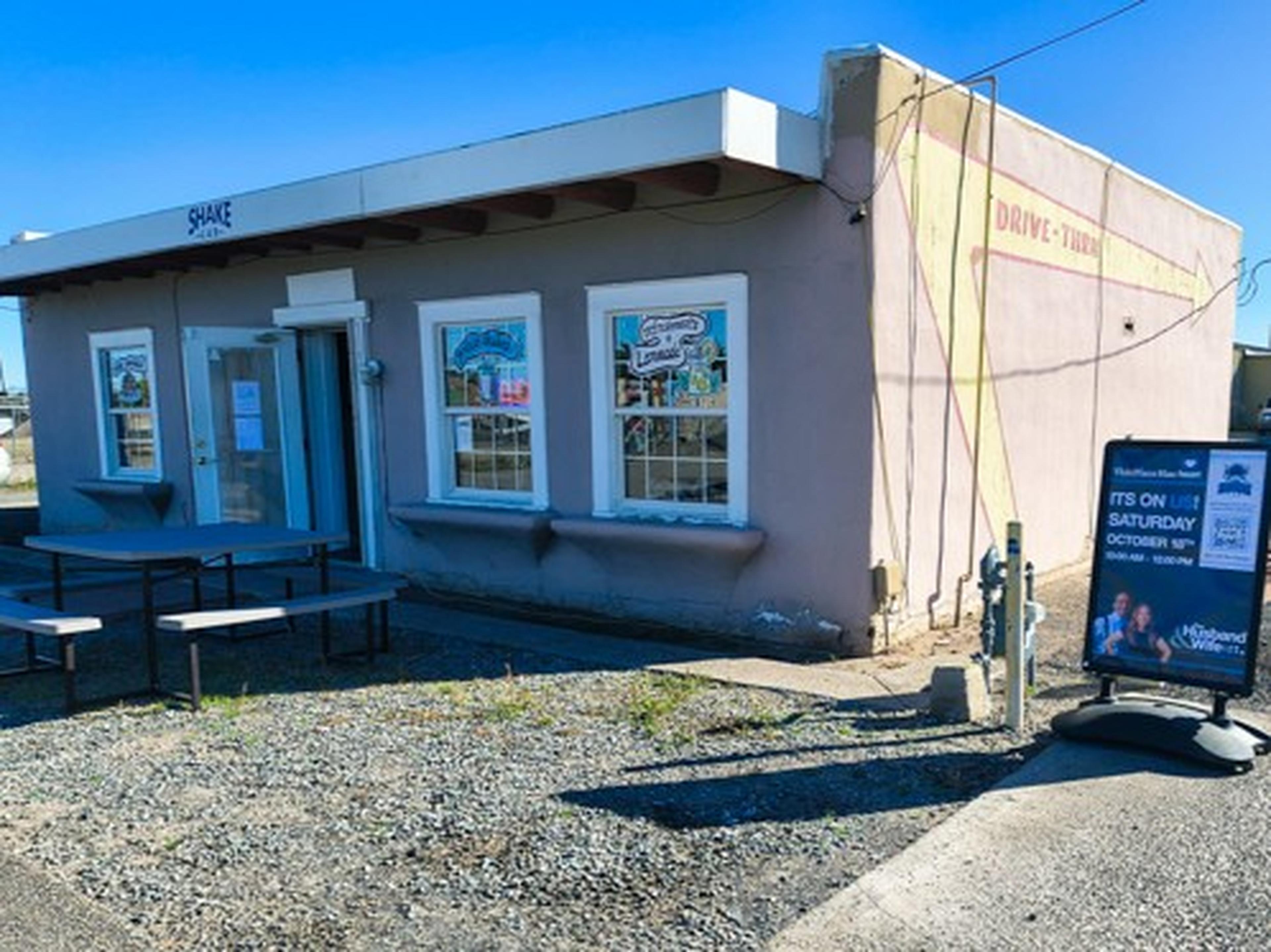 Small beige building with "Shake" sign, drive-thru, outdoor seating, and posters on windows. A signboard advertises an event. Gravel foreground.