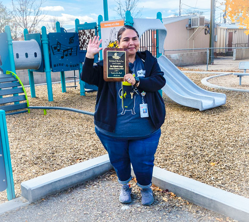Mrs. Lozoya smiles and waves with her new Teacher of the Month plaque Teacher in front of playground holding a plaque and waving with a smile