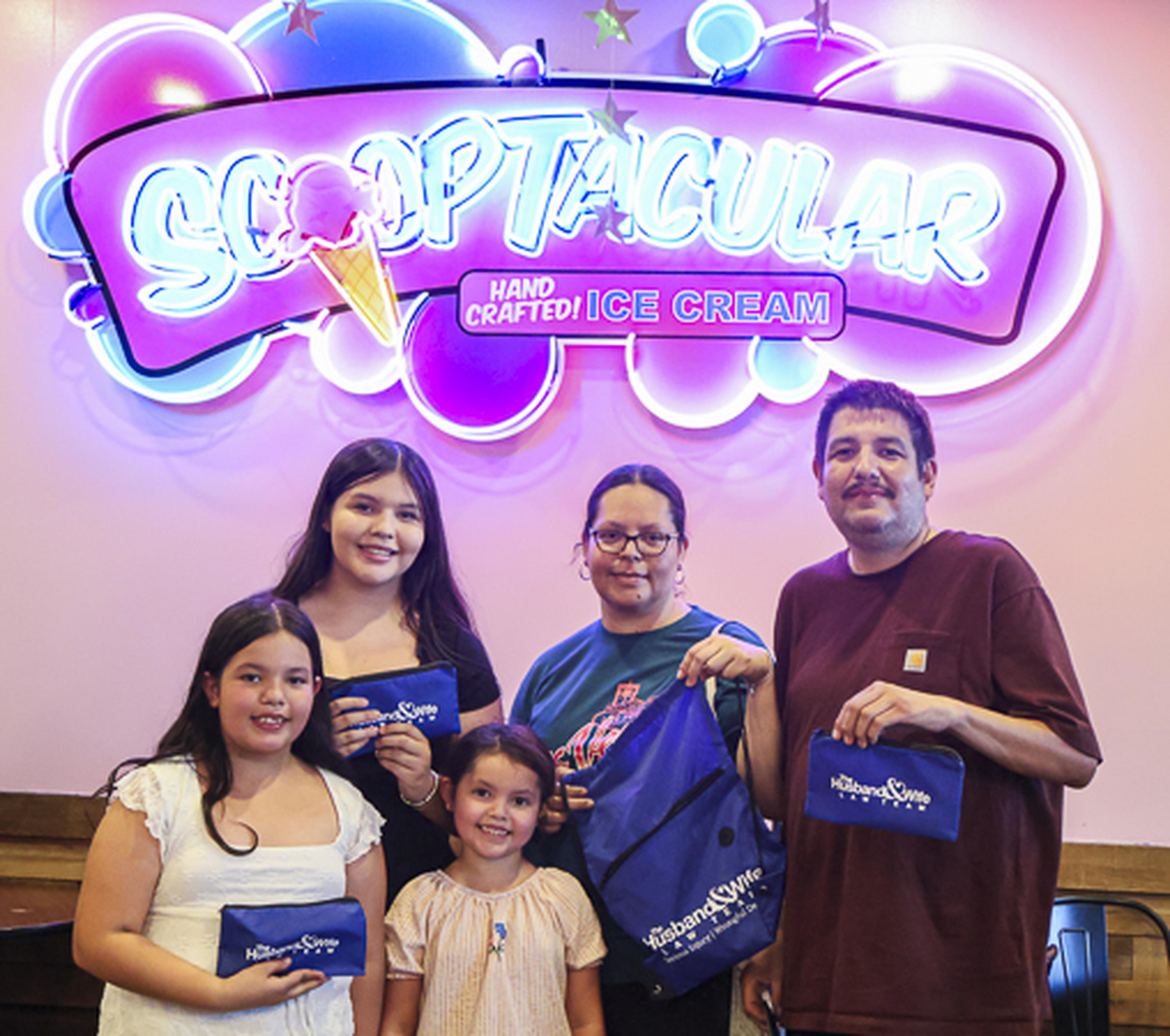 A family of five smiles together in front of the SCOOPTACULAR Ice Cream sign, showing off their Husband & Wife Law Team fans during a fun-filled community event.