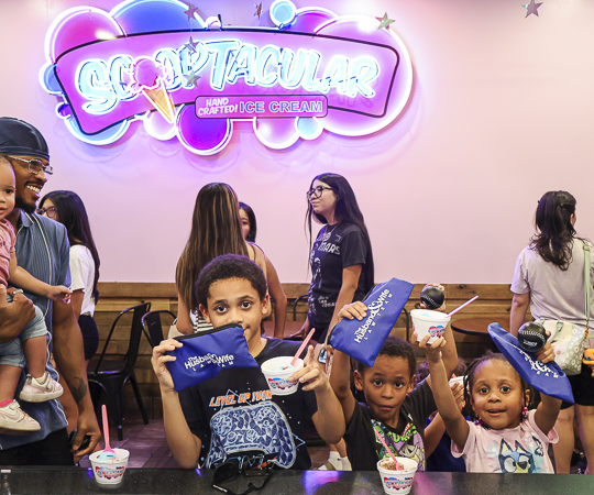 Children enjoy ice cream and Husband & Wife Law Team giveaways at the SCOOPTACULAR HEART Event, making sweet memories together under the bright neon sign.