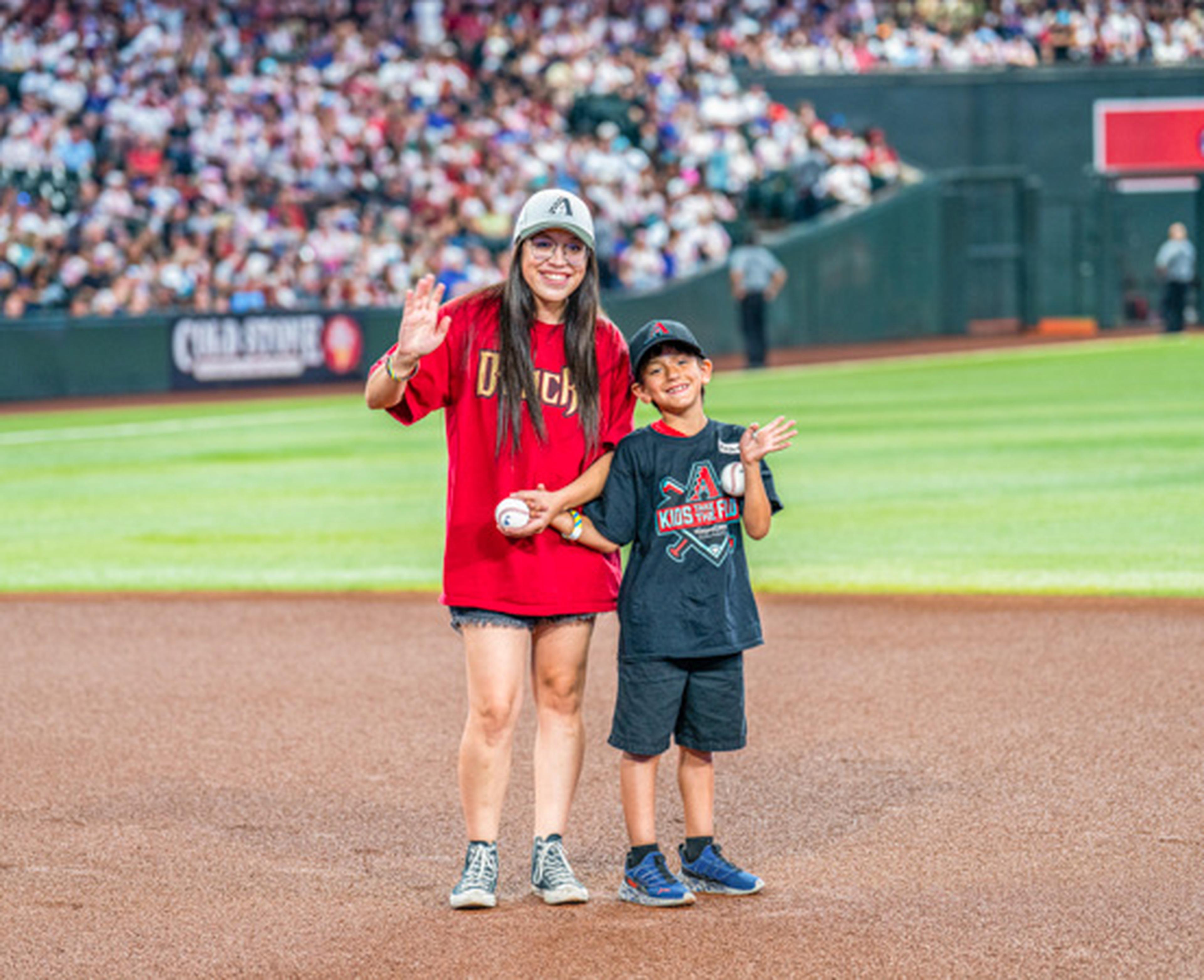 Diamondbacks duo! Mom and son wave from the field in matching team gear, the crowd cheering behind them.