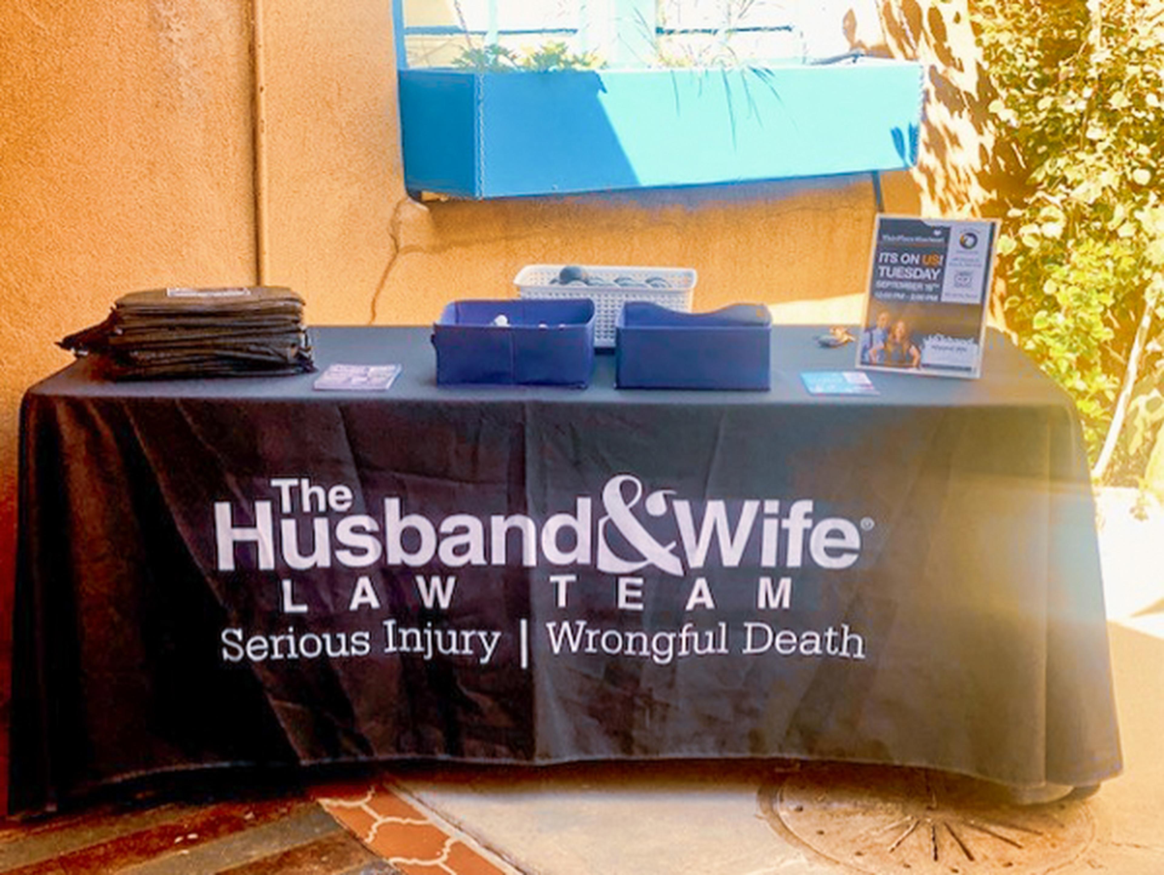 Table with a black cloth displaying "The Husband & Wife Law Team" for serious injury and wrongful death; brochures and bags on top.