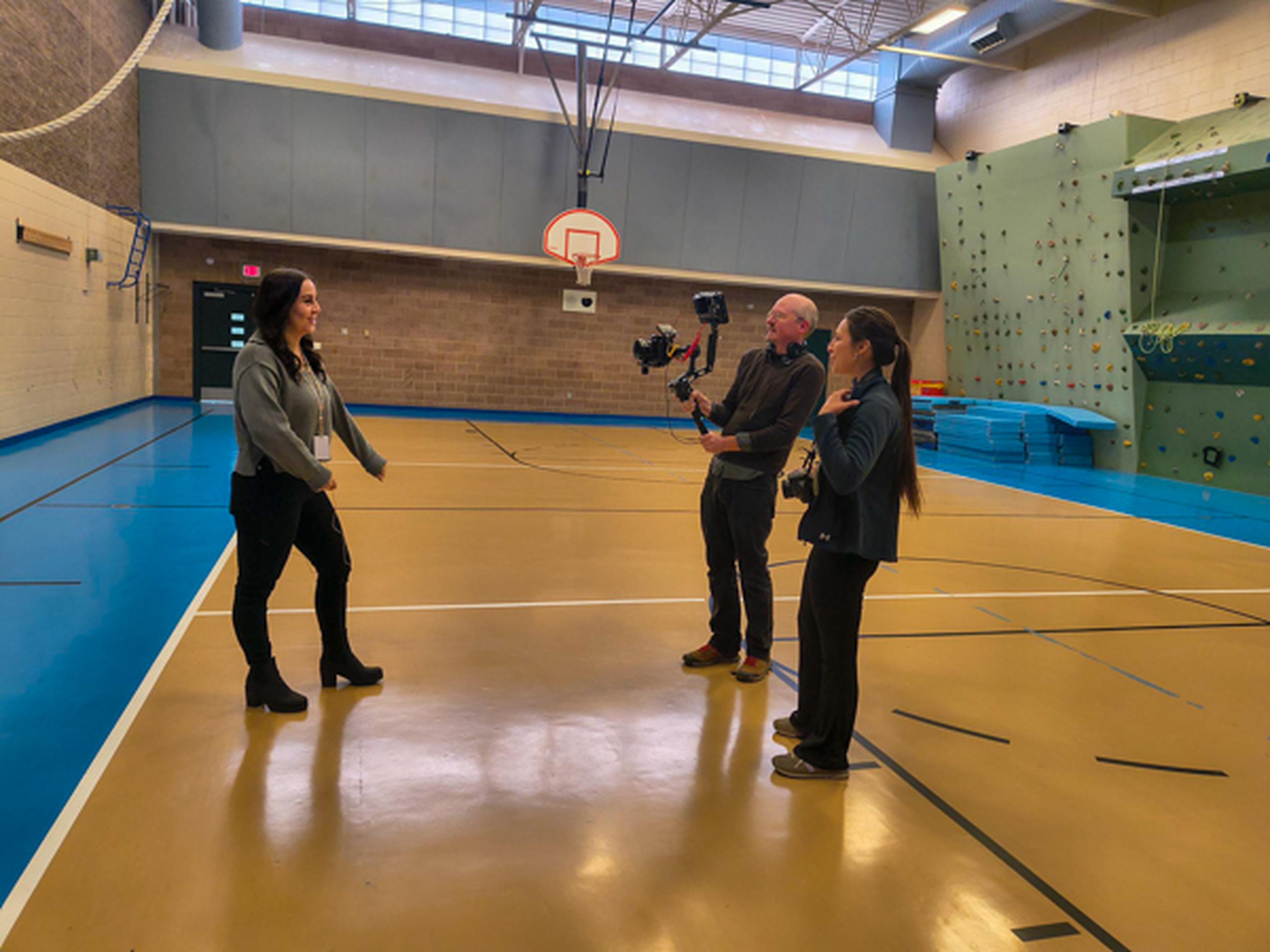 Three people in a gym with filming equipment. One is on-camera, while two operate a camera on a stabilizer. Climbing wall is visible.