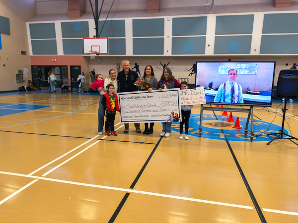 A group of people in a gym holding a large check, standing near a TV screen displaying a person's image. Basketball hoop in the background.