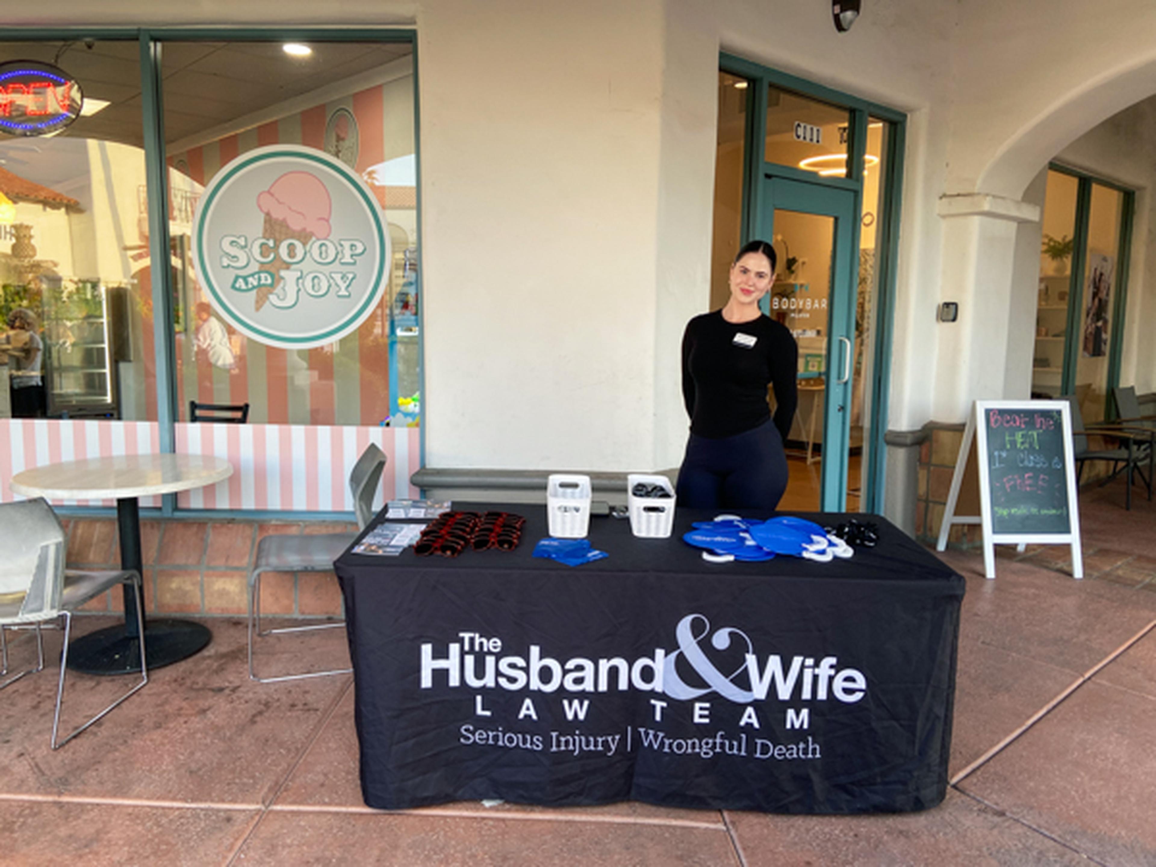 A woman stands behind a table with "The Husband & Wife Law Team" banner, displaying promotional items outside an ice cream shop.