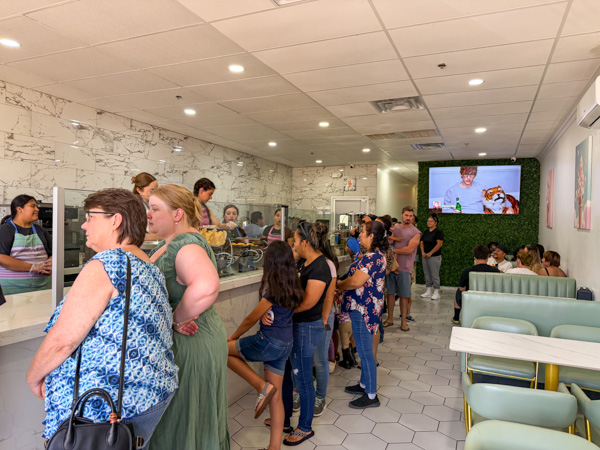 Families and community members in line inside a busy  Iced Out Ice Cream  shop with hexagonal tile floor and a TV on the wall displaying food-related content.