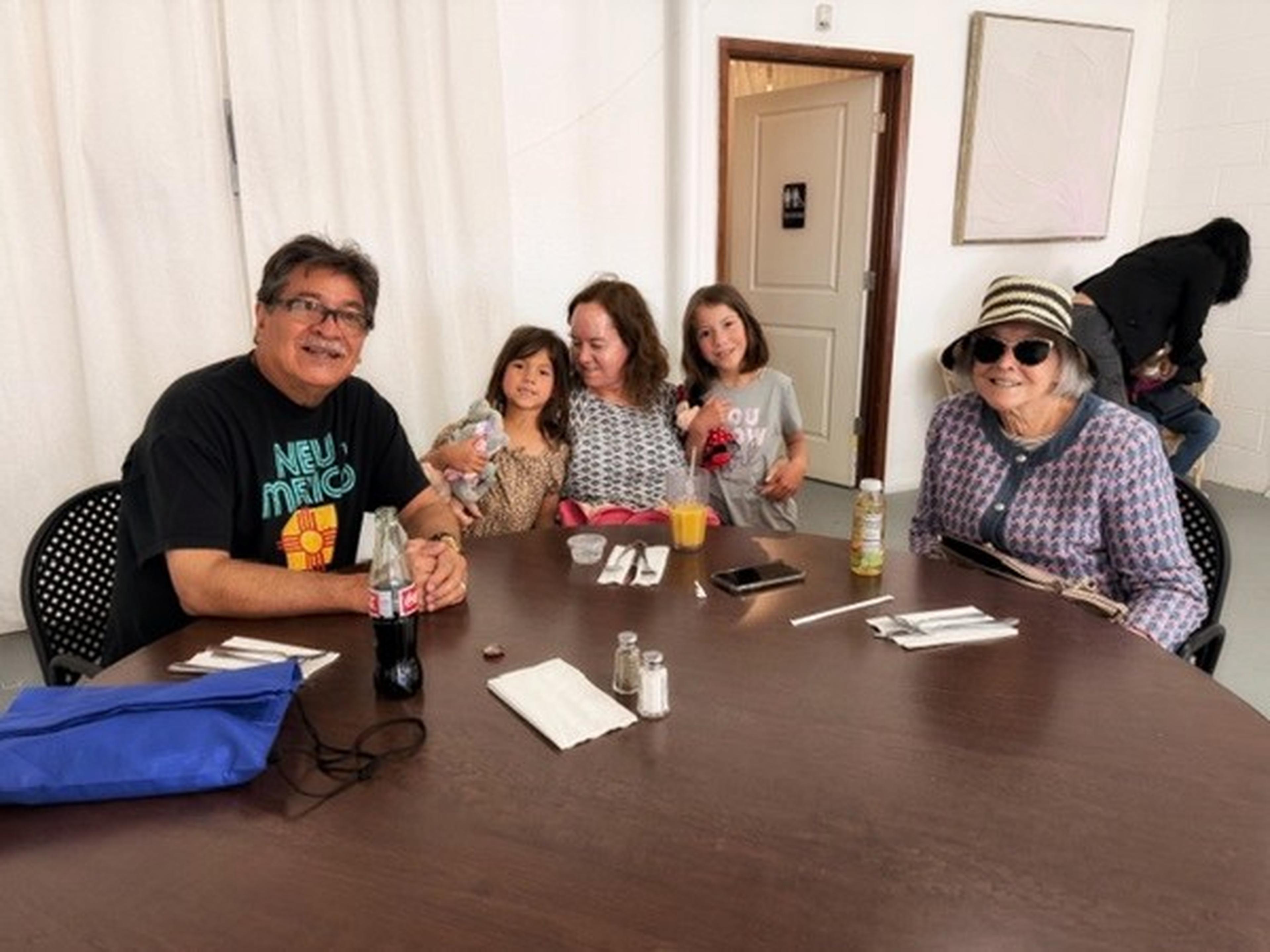 Group of five people sitting around a table with drinks and utensils. The woman on the right wears a sun hat and sunglasses.