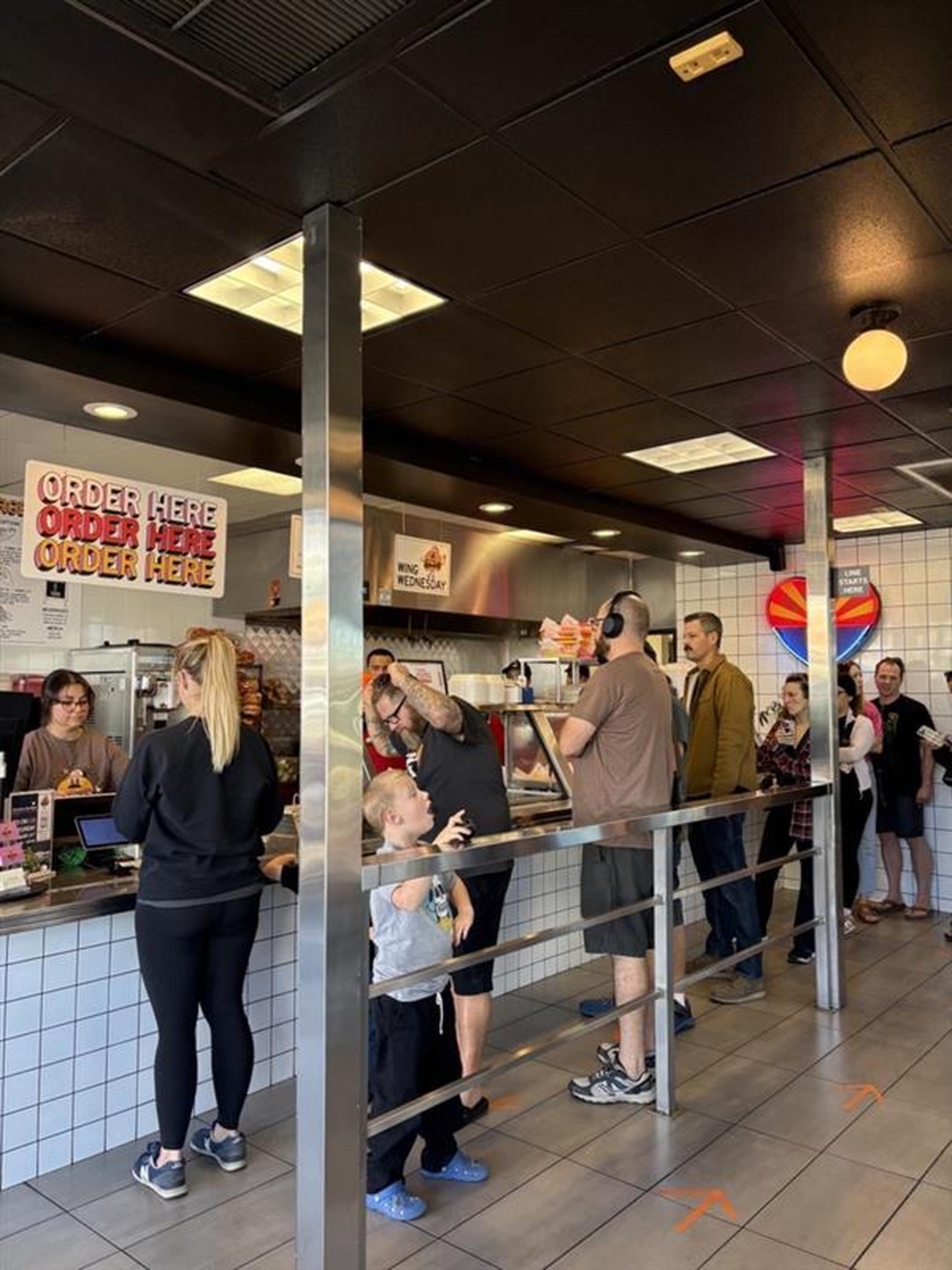 Customers line up inside the main burger location with menu signs on the wall, waiting to order at the counter.