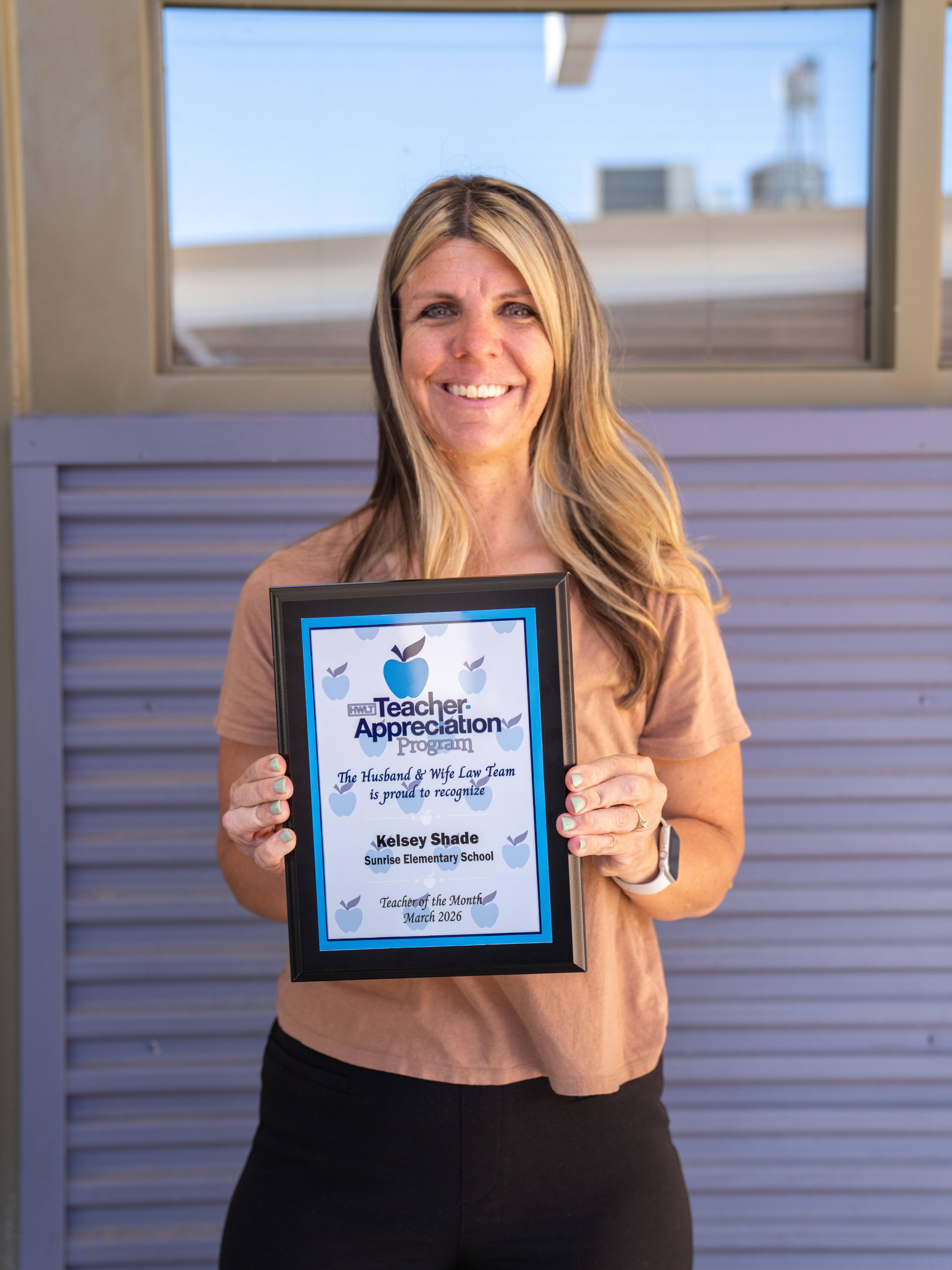 Ms. Shade smiling and holding a framed Teacher Appreciation certificate in front of a window.