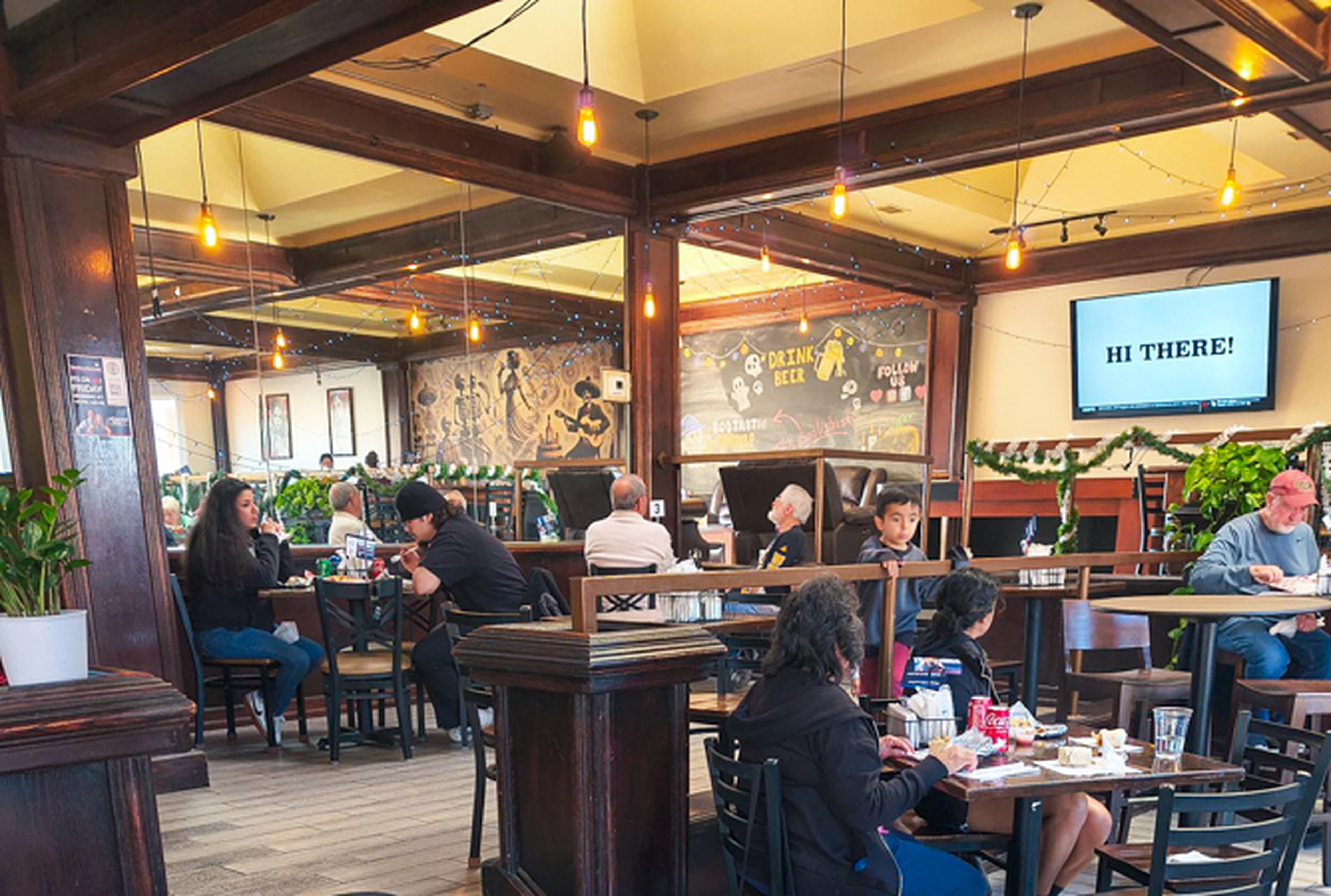 People dining in a cozy restaurant with wooden decor, a mural on the wall, and a TV displaying "Hi There!" in the background.