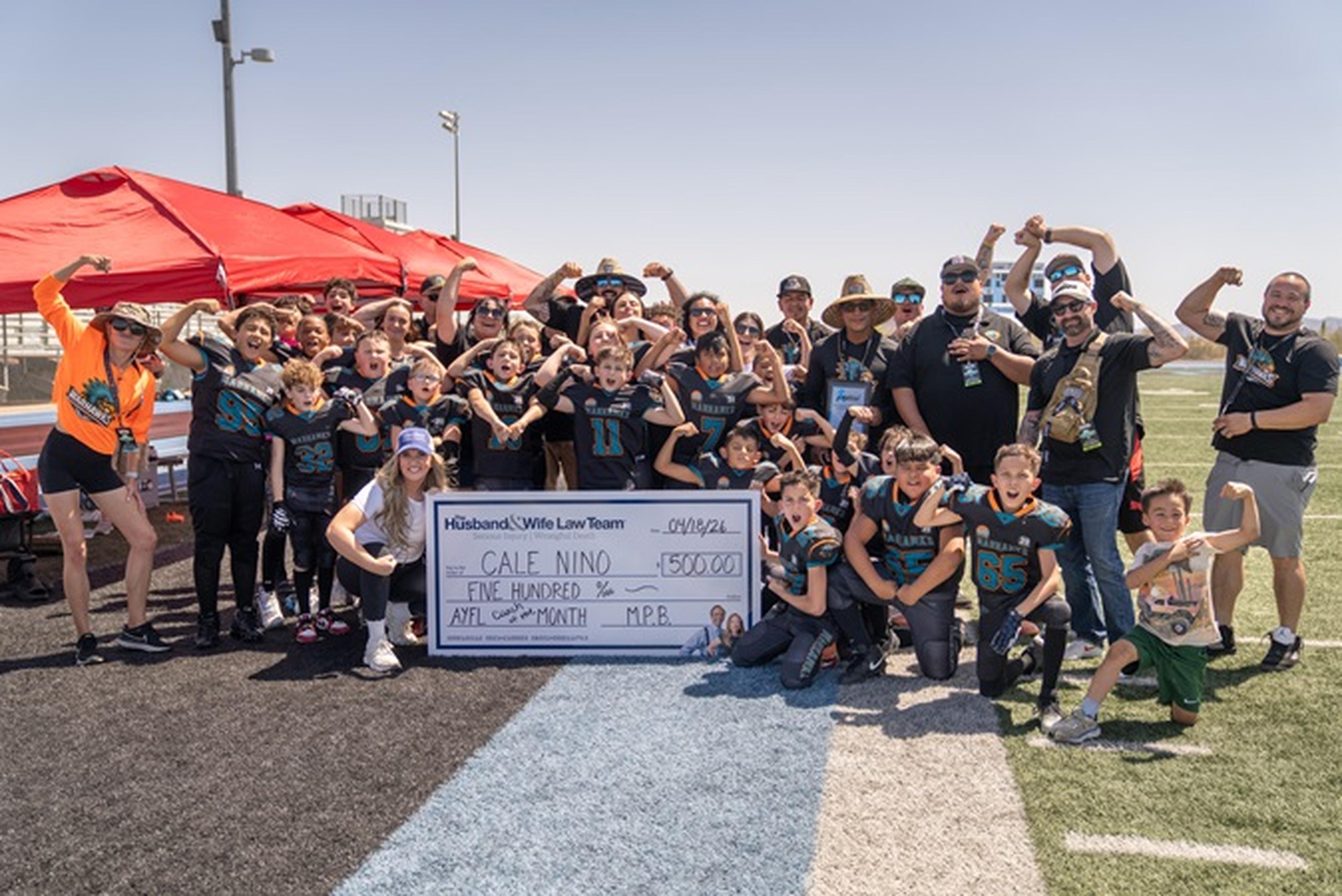 On the football field, AYFL athletes and supporters cheer for Coach Nino as he’s presented with a large check from the Husband & Wife Law Team.