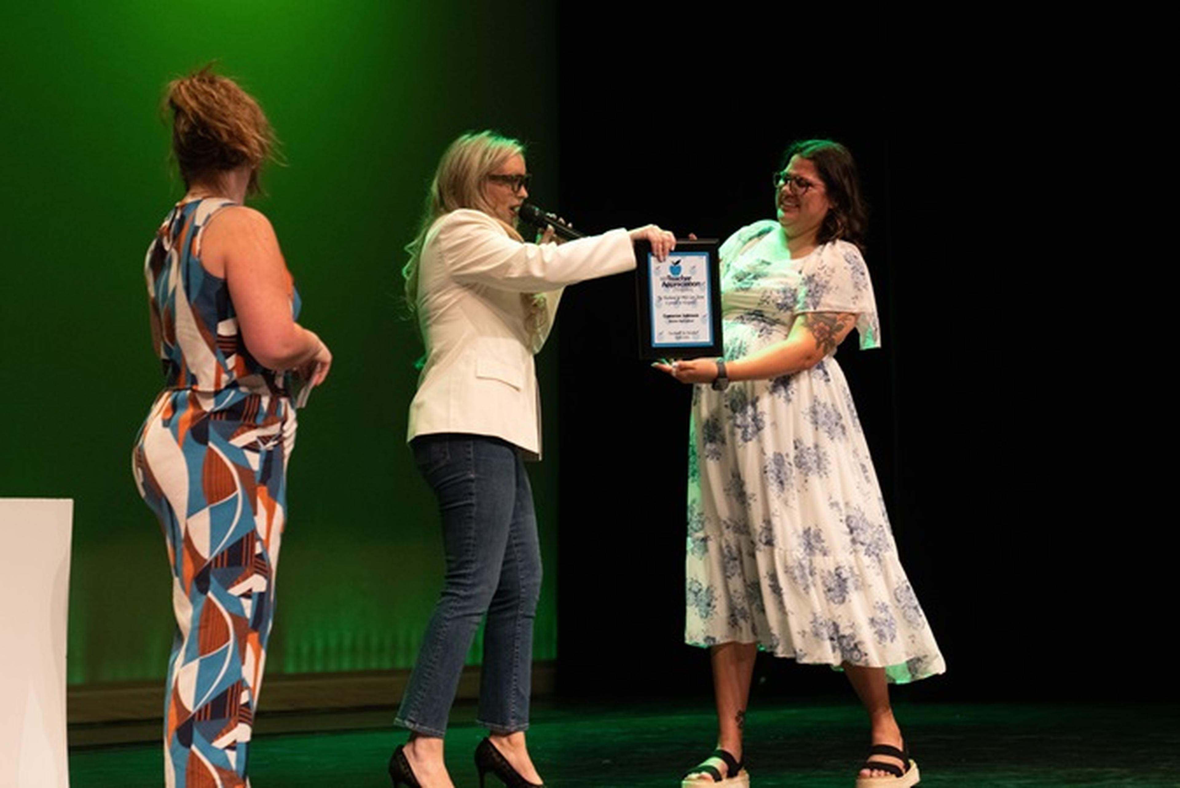 Ms. Johnson accepts a framed Husband and Wife Teacher Appreciation Award, standing before a backdrop glowing green.