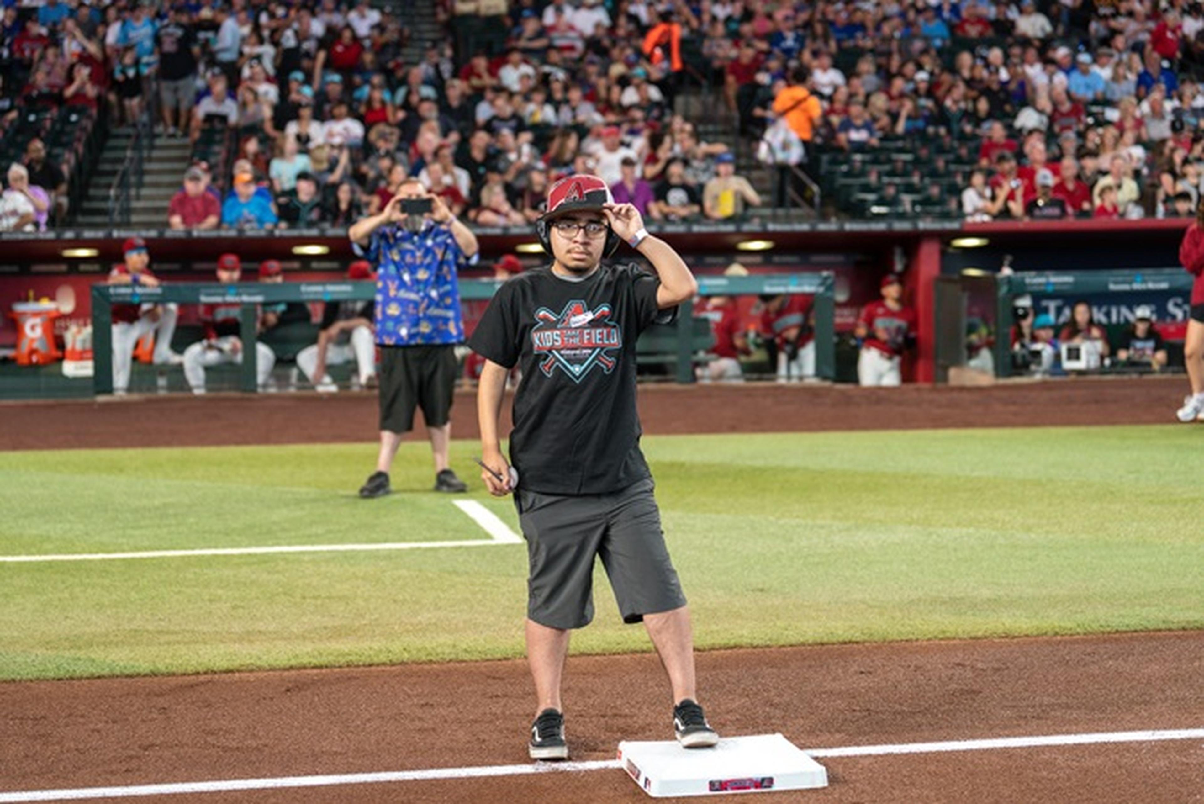 Person in a black t-shirt and cap stands on a baseball field near a base, with a person taking photos and a crowd in the background.