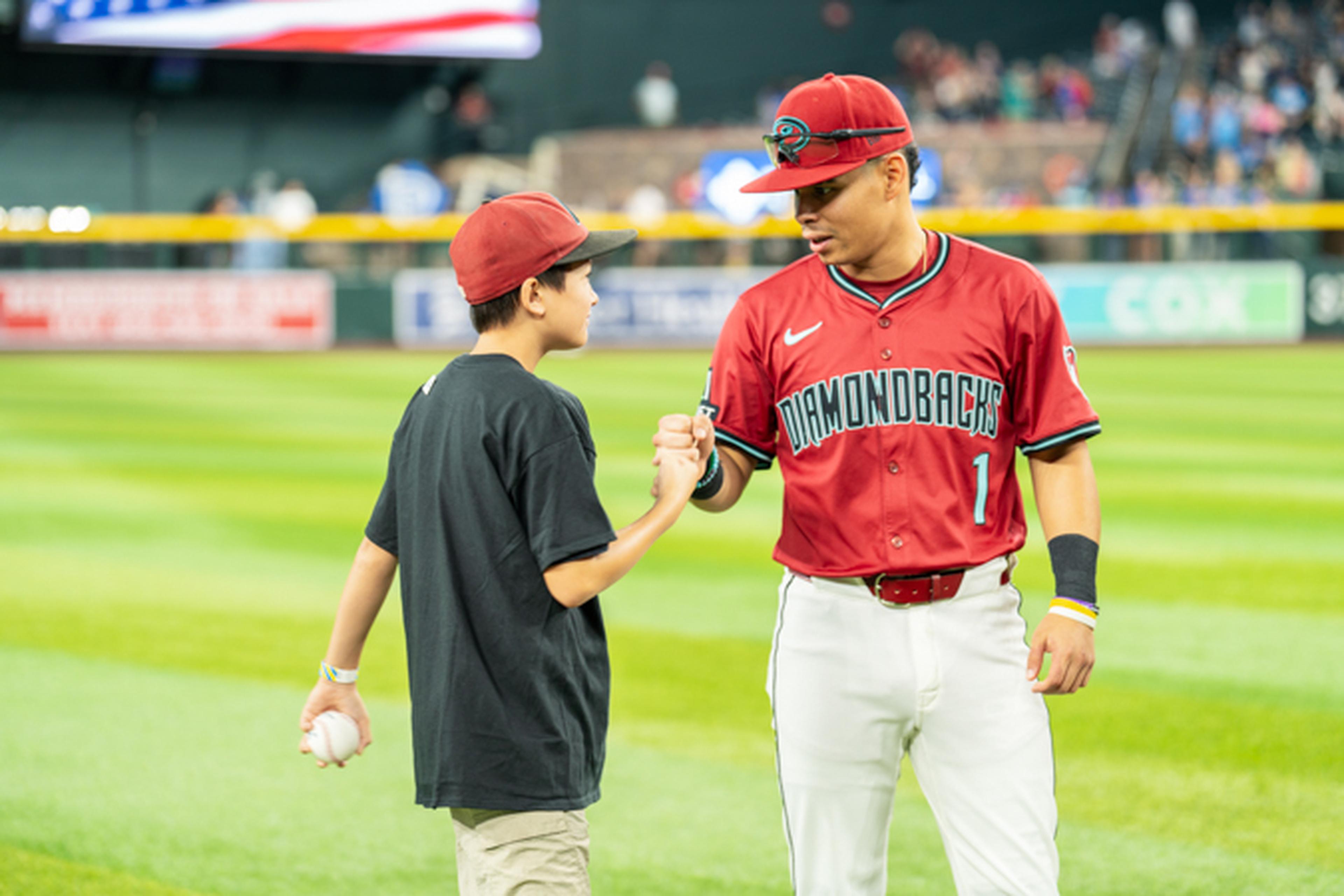 A baseball player in a red jersey fist-bumps a young fan holding a baseball on a field, with spectators in the background.
