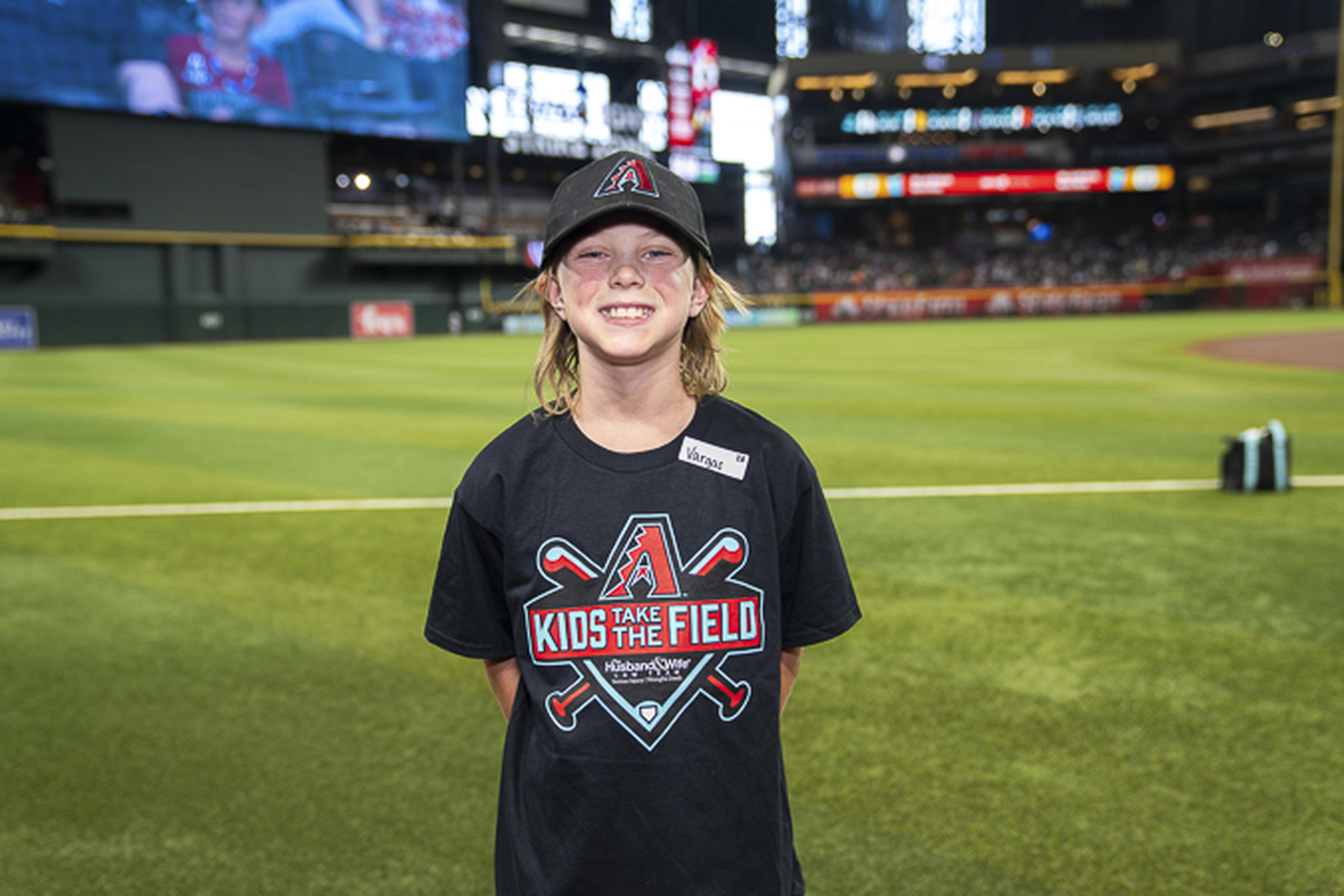 Smiling on the stadium turf, a child wearing a “Kids Take the Field” tee and baseball cap stands in front of the bustling crowd and scoreboard.