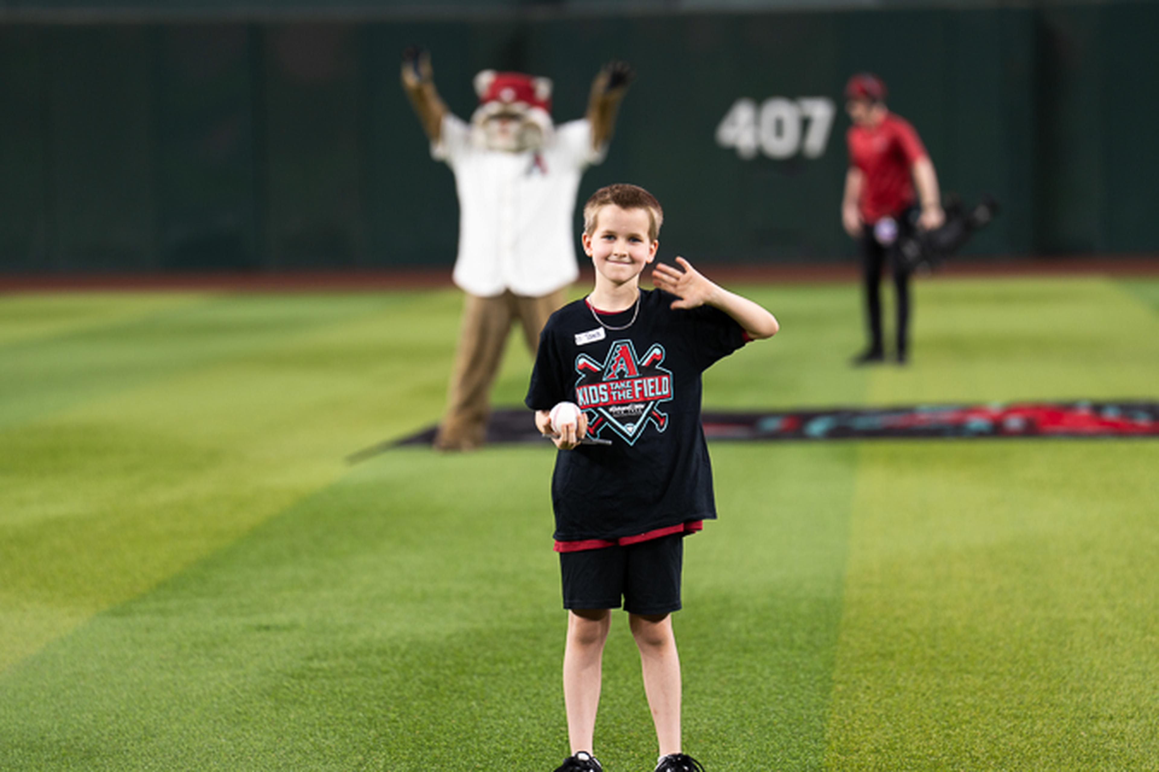 A young Kids Take the Field participant waves from the field while enjoying a memorable experience at Chase Field, with Baxter and fellow game-day activities in the background