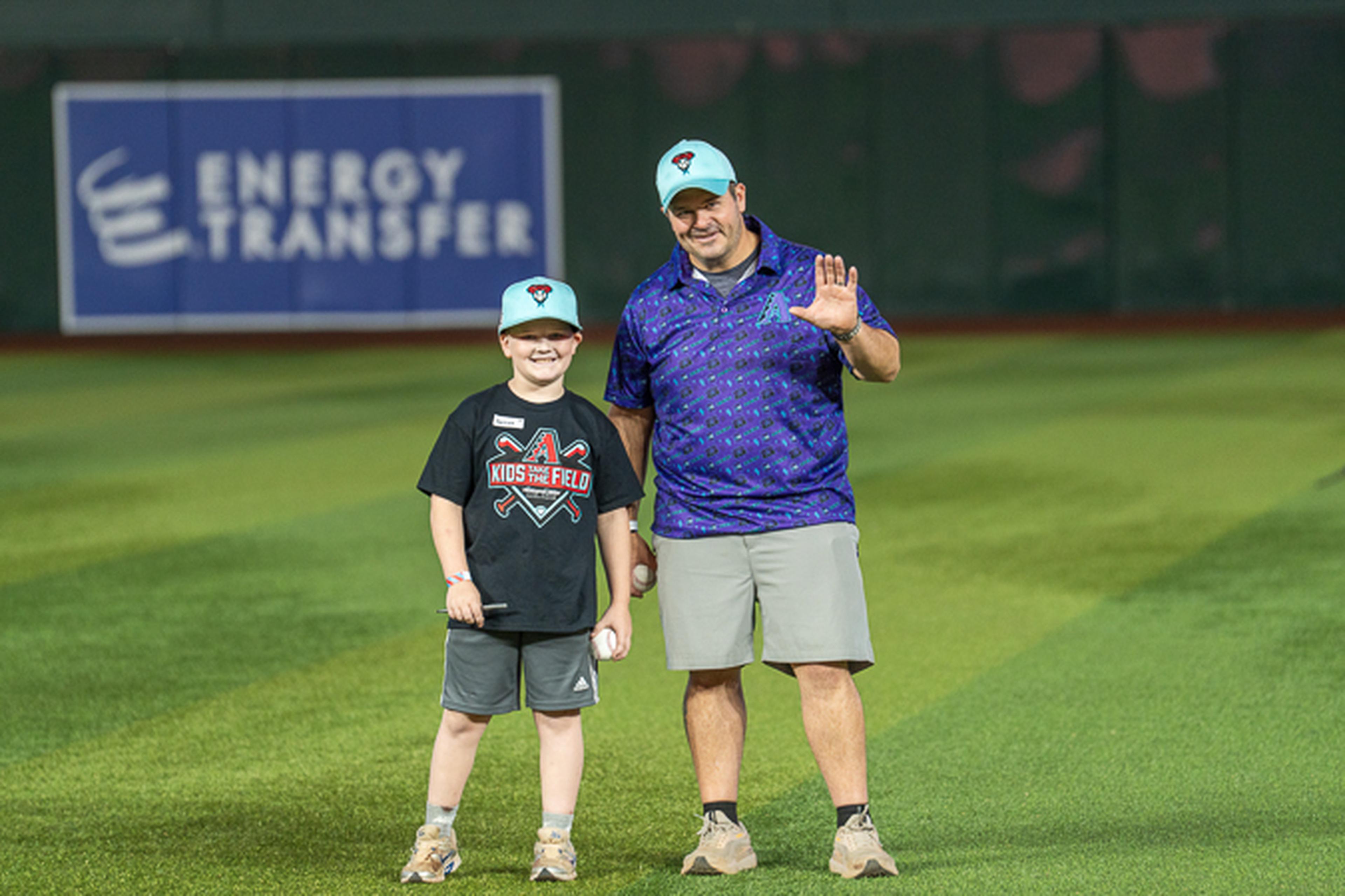 Father and son wearing matching Diamondbacks caps stand smiling on the baseball field, with an “Energy Transfer” sign visible in the background.
