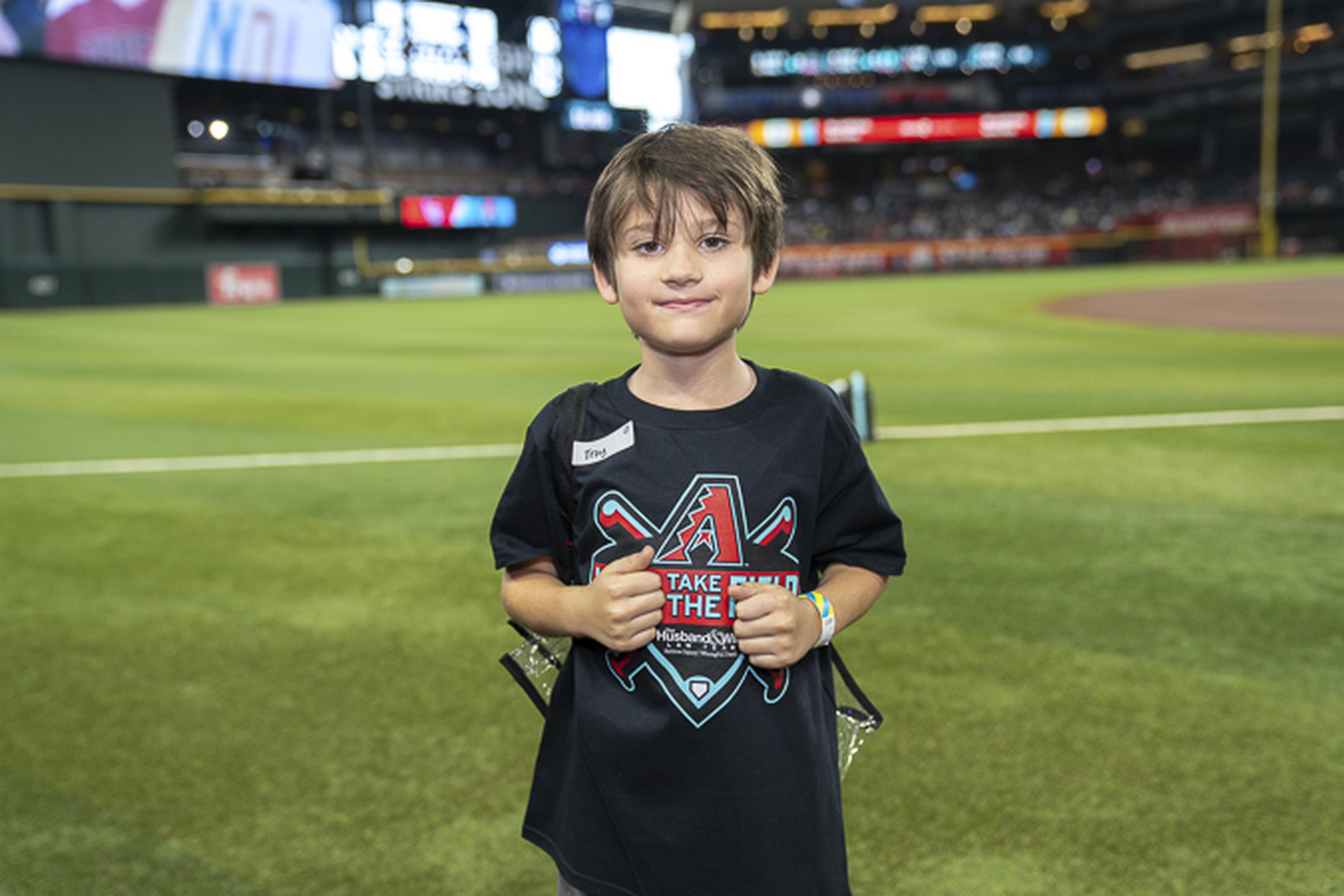 A young boy in a black baseball‑themed T‑shirt smiles on the stadium field with cheering fans in the background.