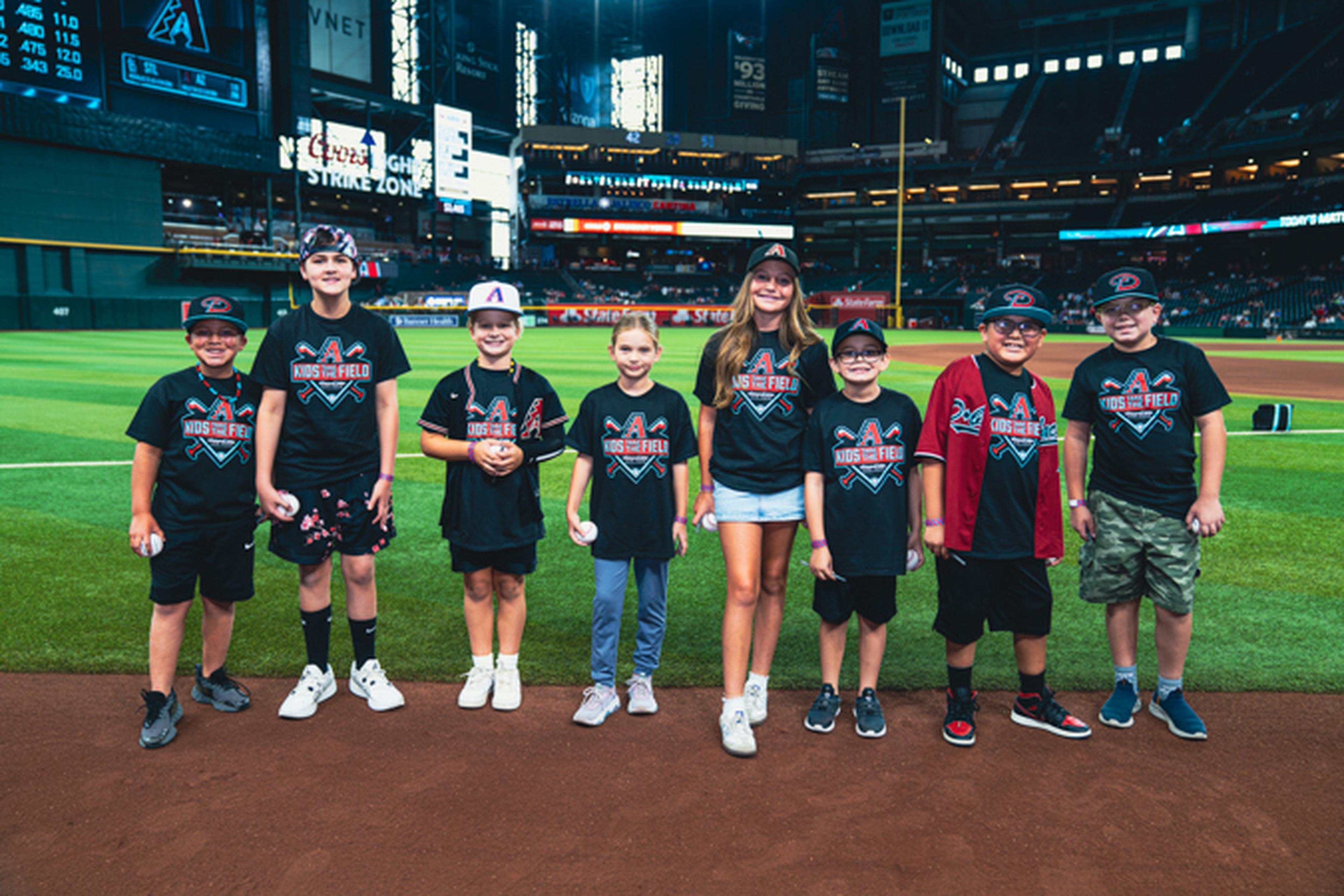 A group of children wearing matching baseball-themed shirts and caps stand on a baseball field, smiling for a photo.